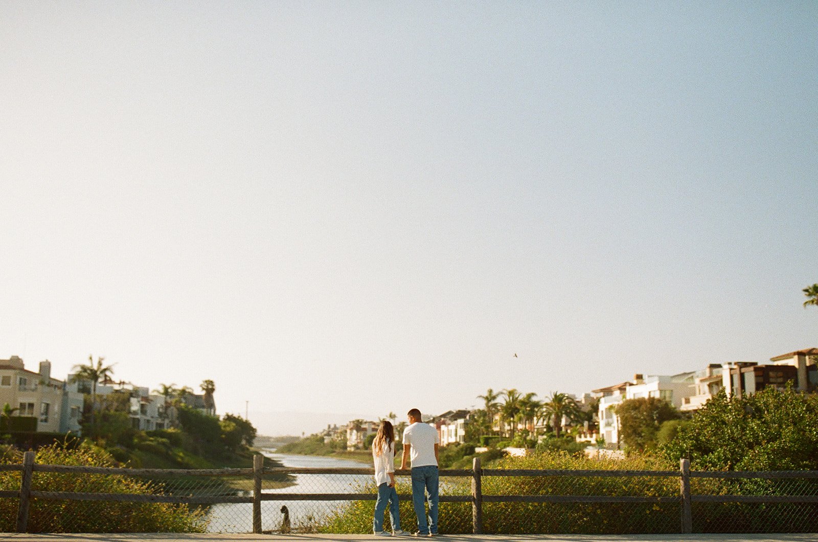 A wide coastal view of a couple standing side by side near Venice Beach Photoshoot Locations with palm trees and calm water behind them.