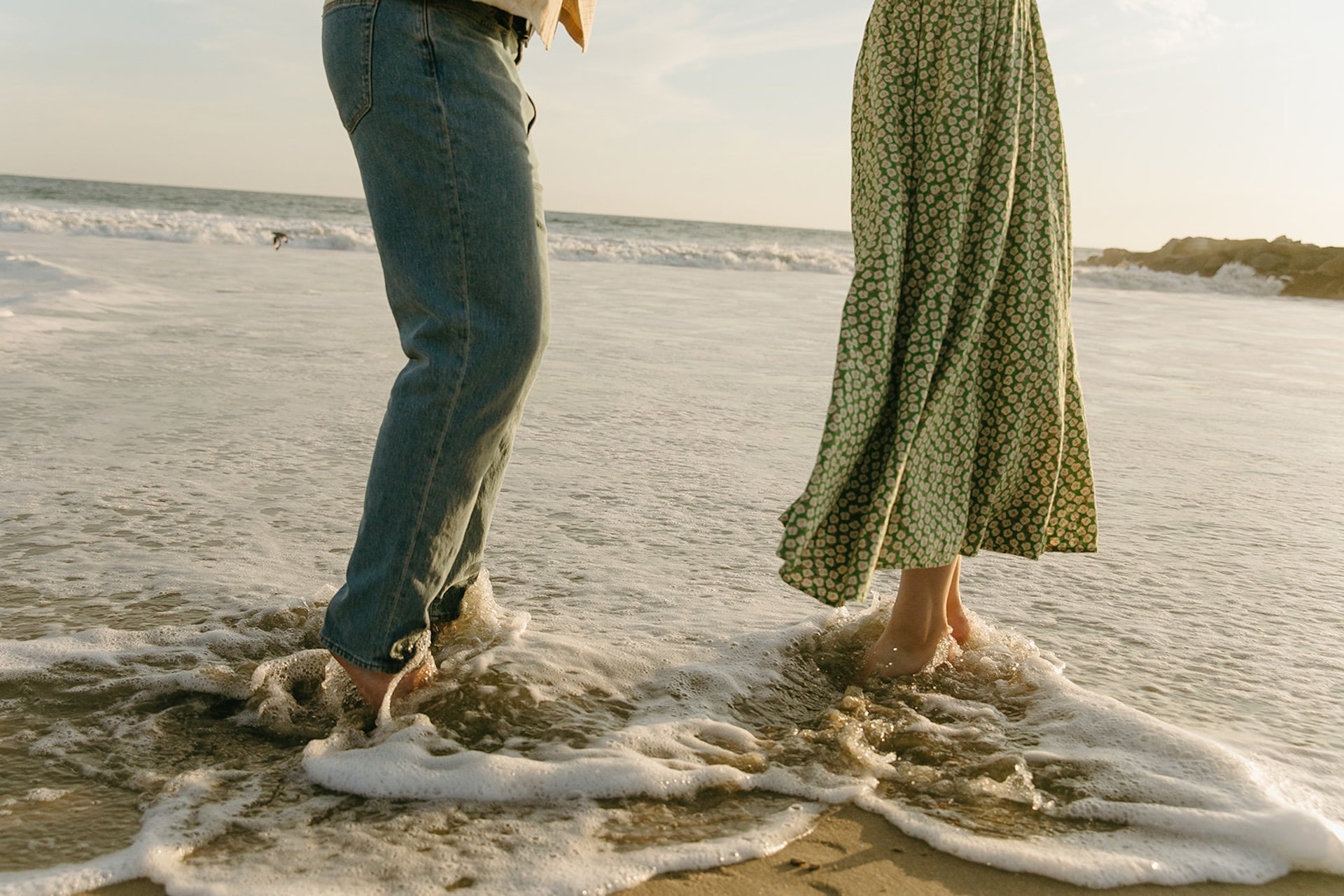 A detail photo of a couples feet walking on the sand as the waves crash against their feet.