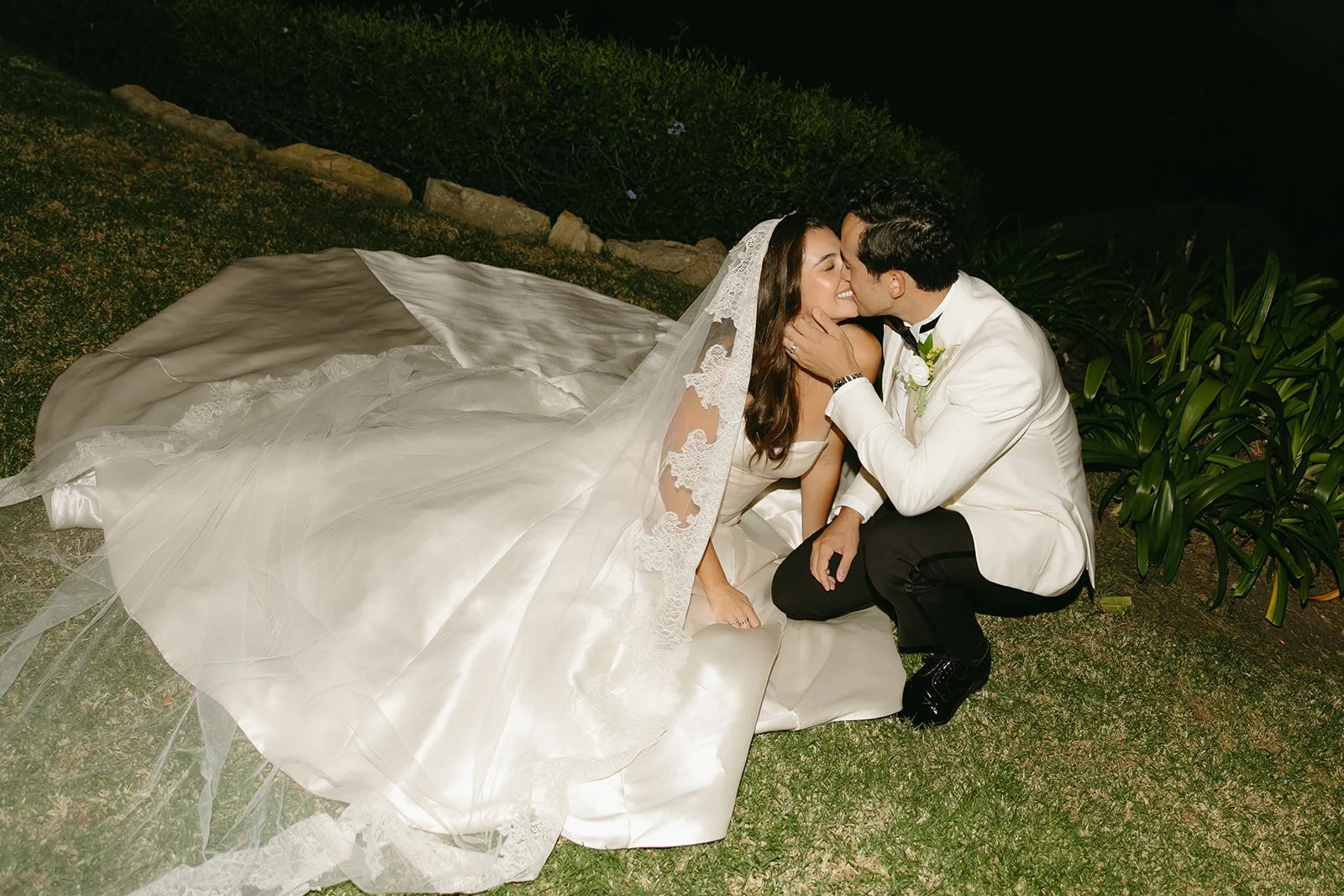 Bride and groom sitting together on the grass sharing a kiss during nighttime portraits at a Palos Verdes wedding venue.