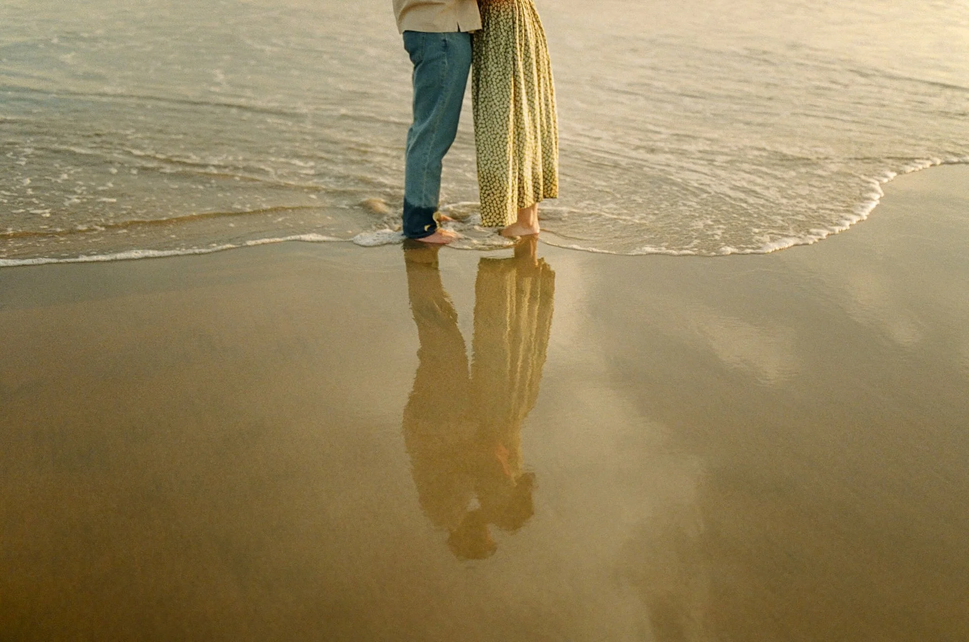 An artistic photo of a couple standing and kissing on the beach with their reflection in the sand.