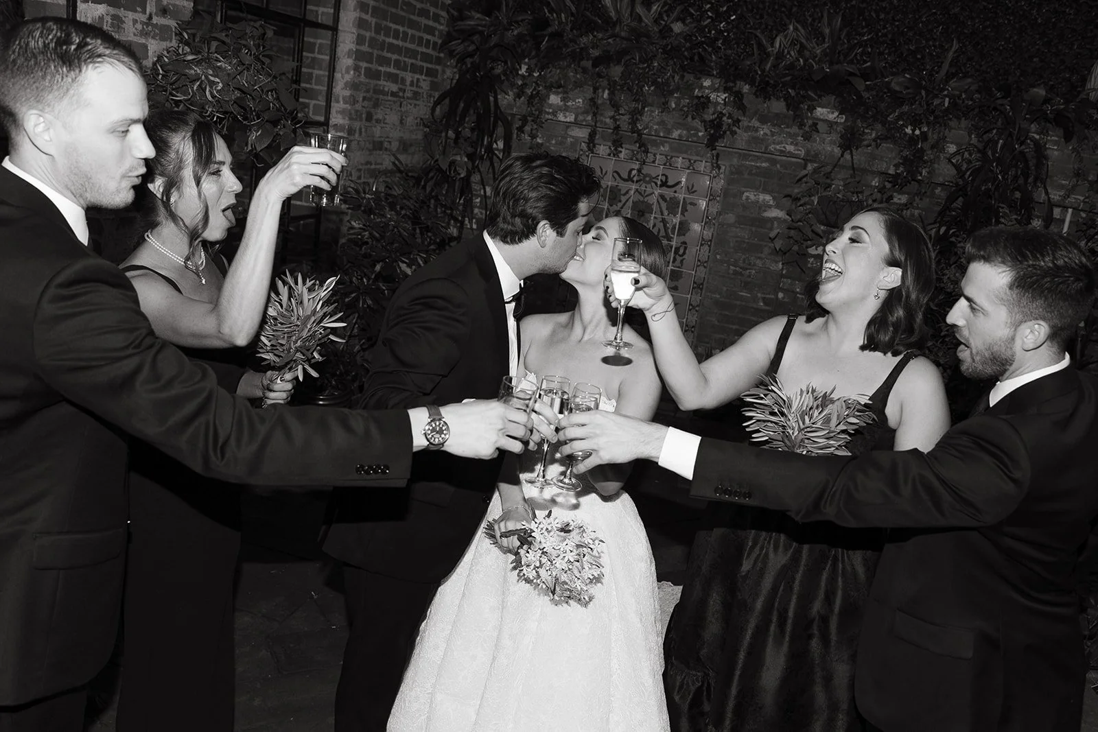 A black and white photo of the wedding party celebrating around the bride and groom as they share a kiss