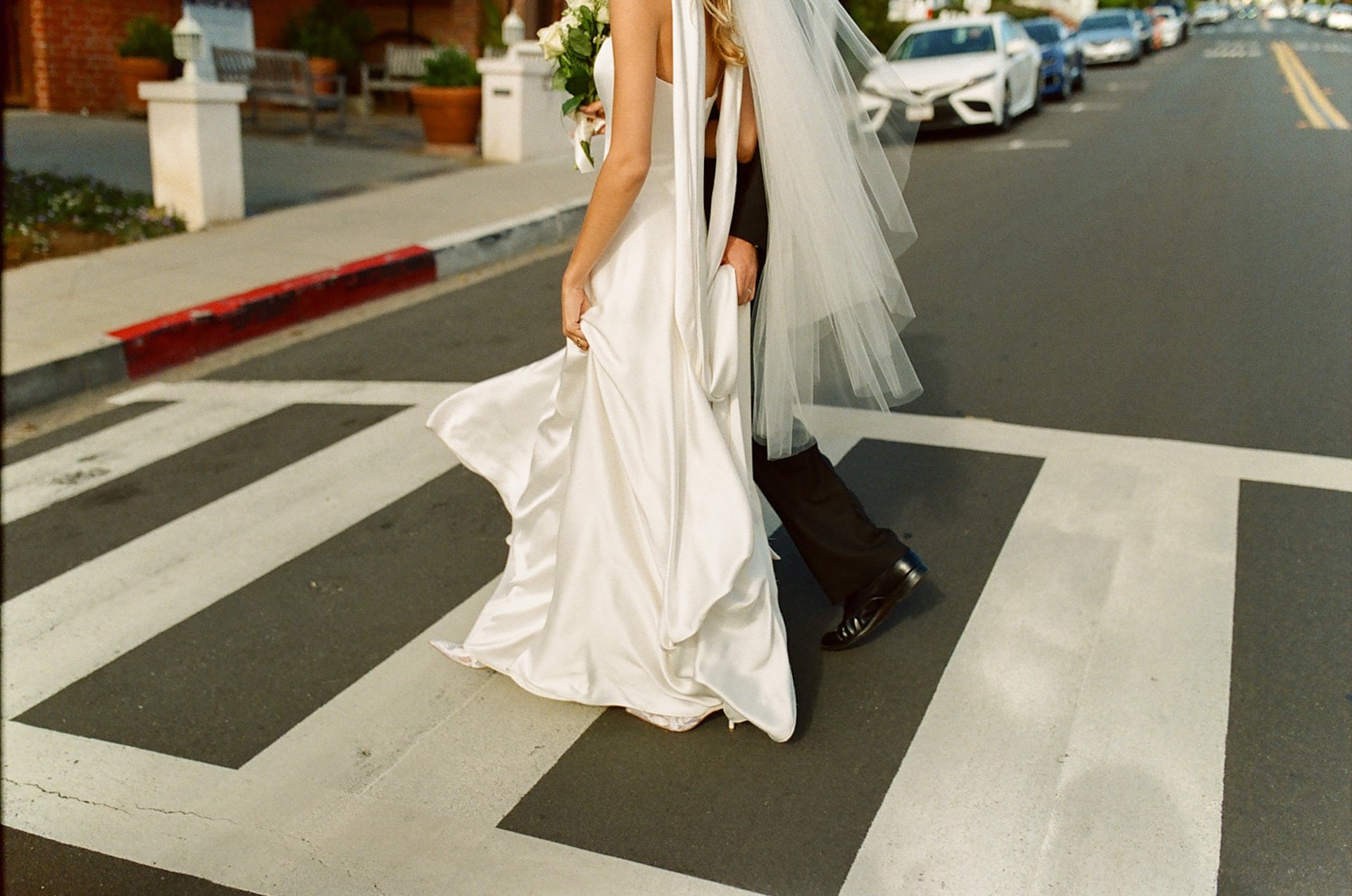 Bride and groom crossing a Laguna Beach street together after their Laguna Beach wedding ceremony, with the bride lifting her satin gown and veil.