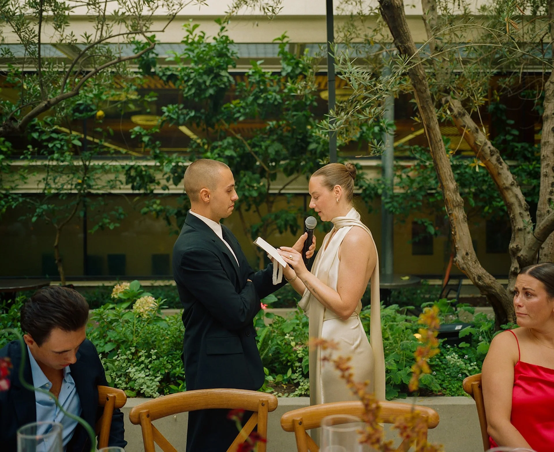 The bride reading her vows into a microphone while standing across from the groom in a lush courtyard setting with guests seated nearby.