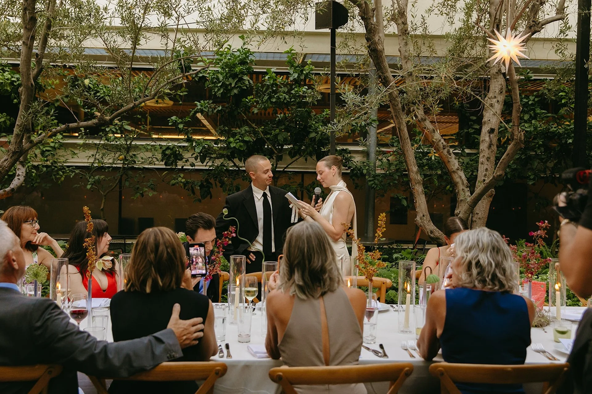 Guests seated at a long outdoor dinner table during a garden wedding reception, with candles, floral centerpieces, and the couple speaking in the background.