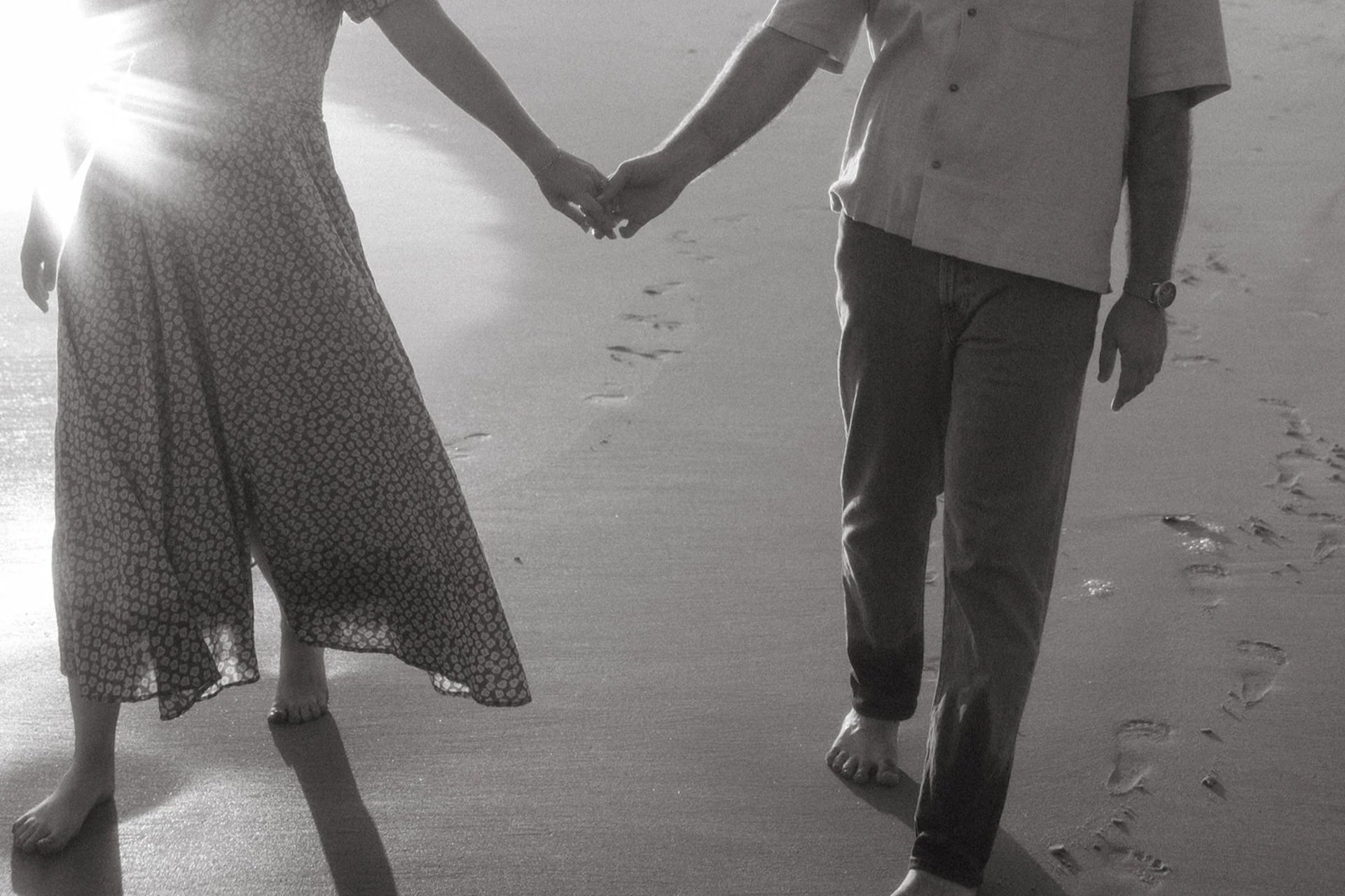 Couple holding hands barefoot in the wet sand at the shoreline, their reflections glowing in the warm evening light.