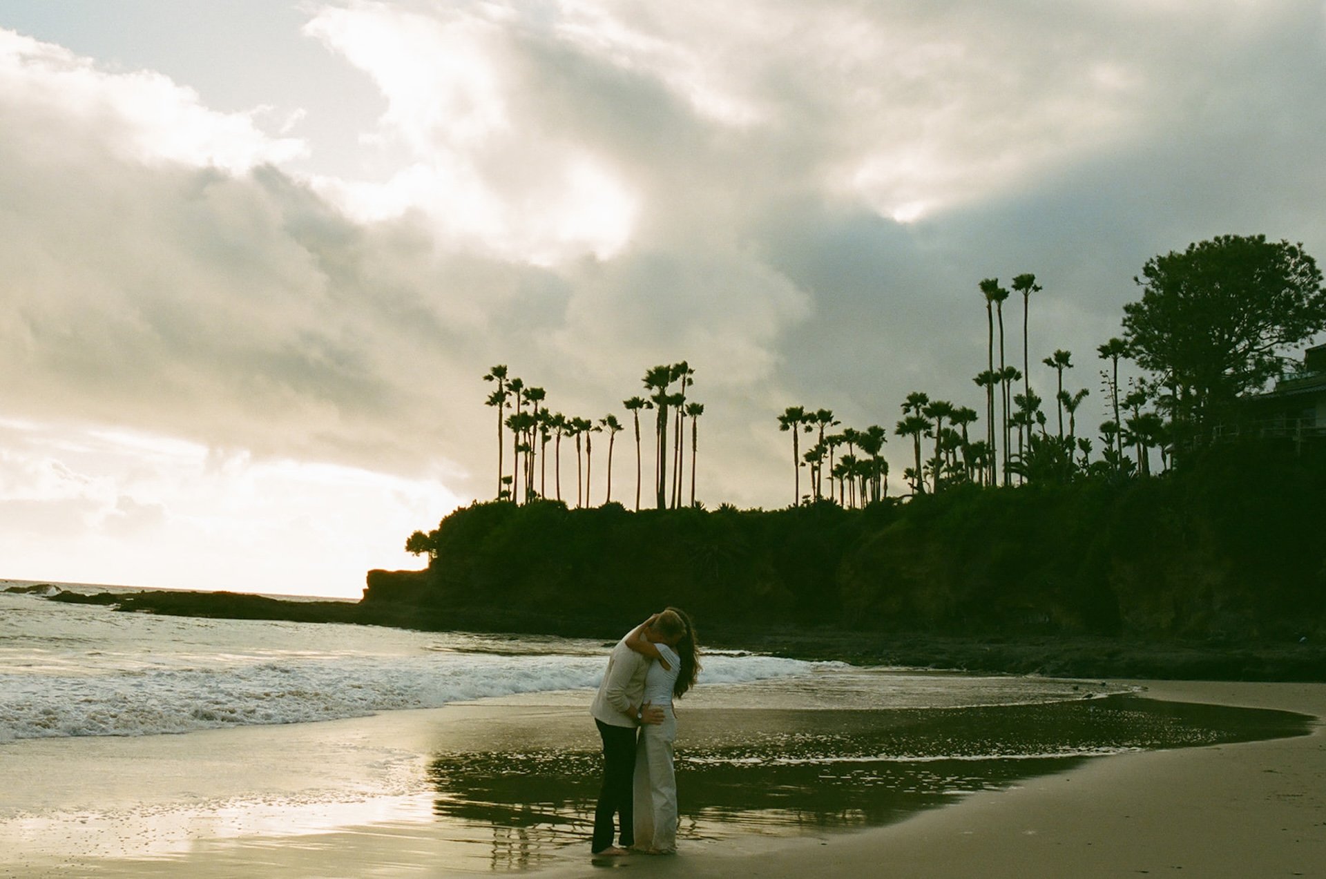 Wide coastal engagement photo of a couple standing together near the water with palm trees and dramatic clouds in the background, calm and romantic beach setting.