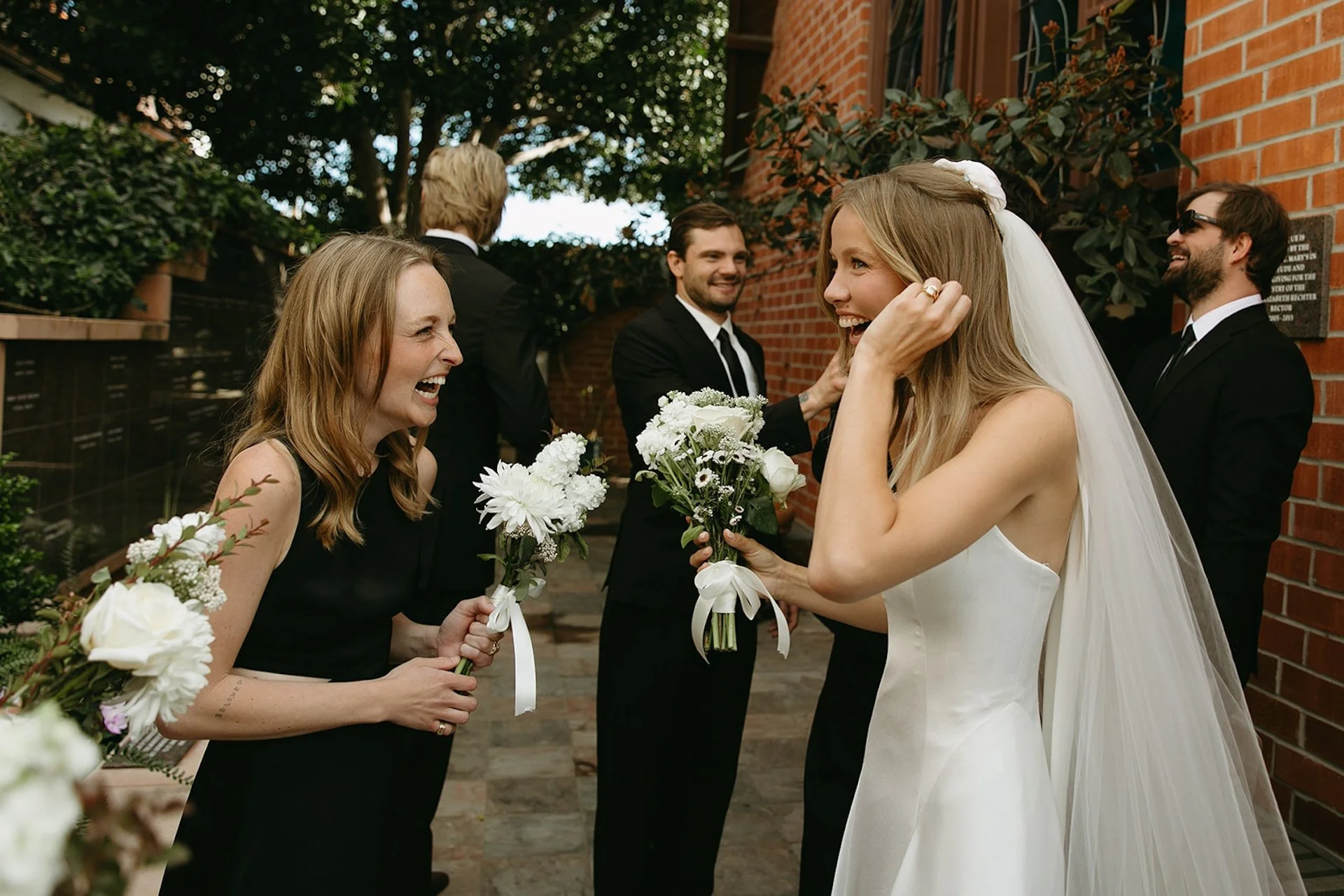 Bride laughing and talking with friends while holding a bouquet beside a brick church courtyard.