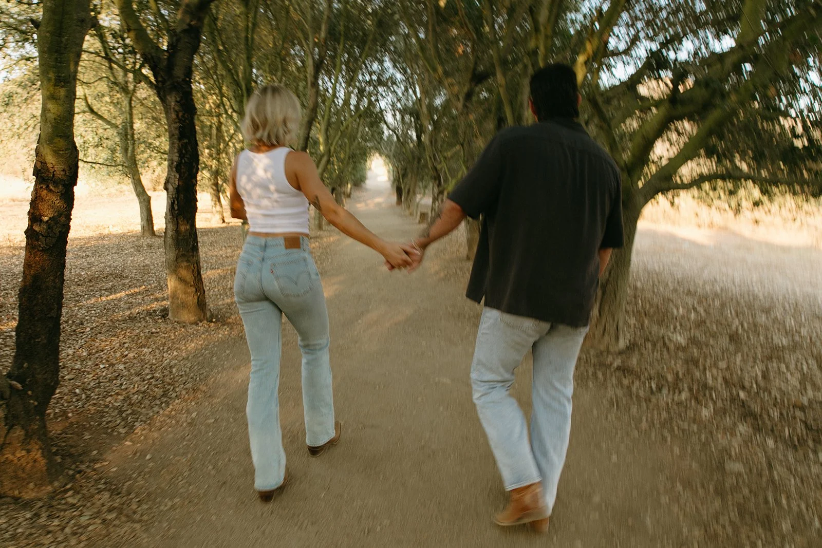 Rear view of the couple holding hands while walking down a tree-lined trail surrounded by warm evening light.