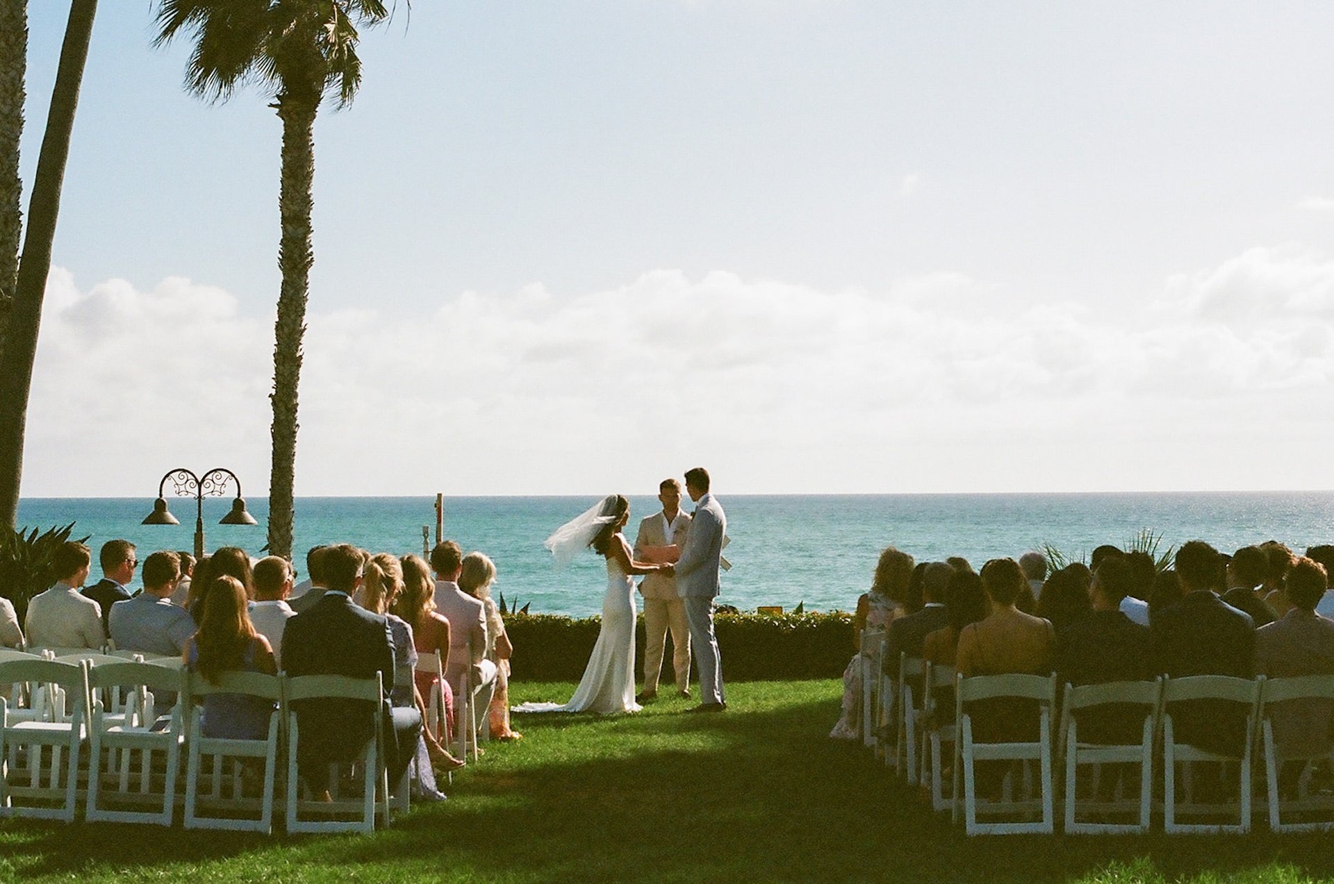 The couple exchanges vows facing the ocean, framed by tall palms and glowing afternoon light.
