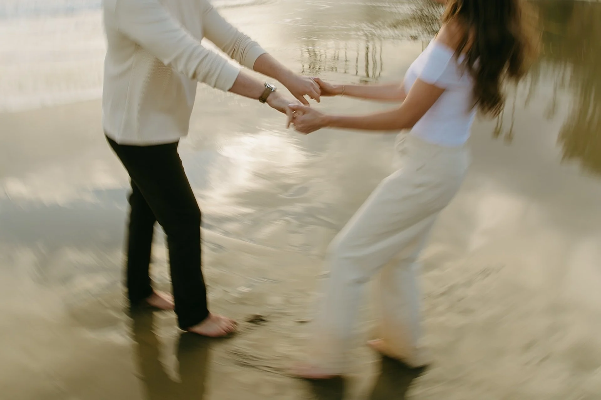 A couple holding hands and spinning playfully in shallow water, motion blur capturing movement and reflections in the wet sand.