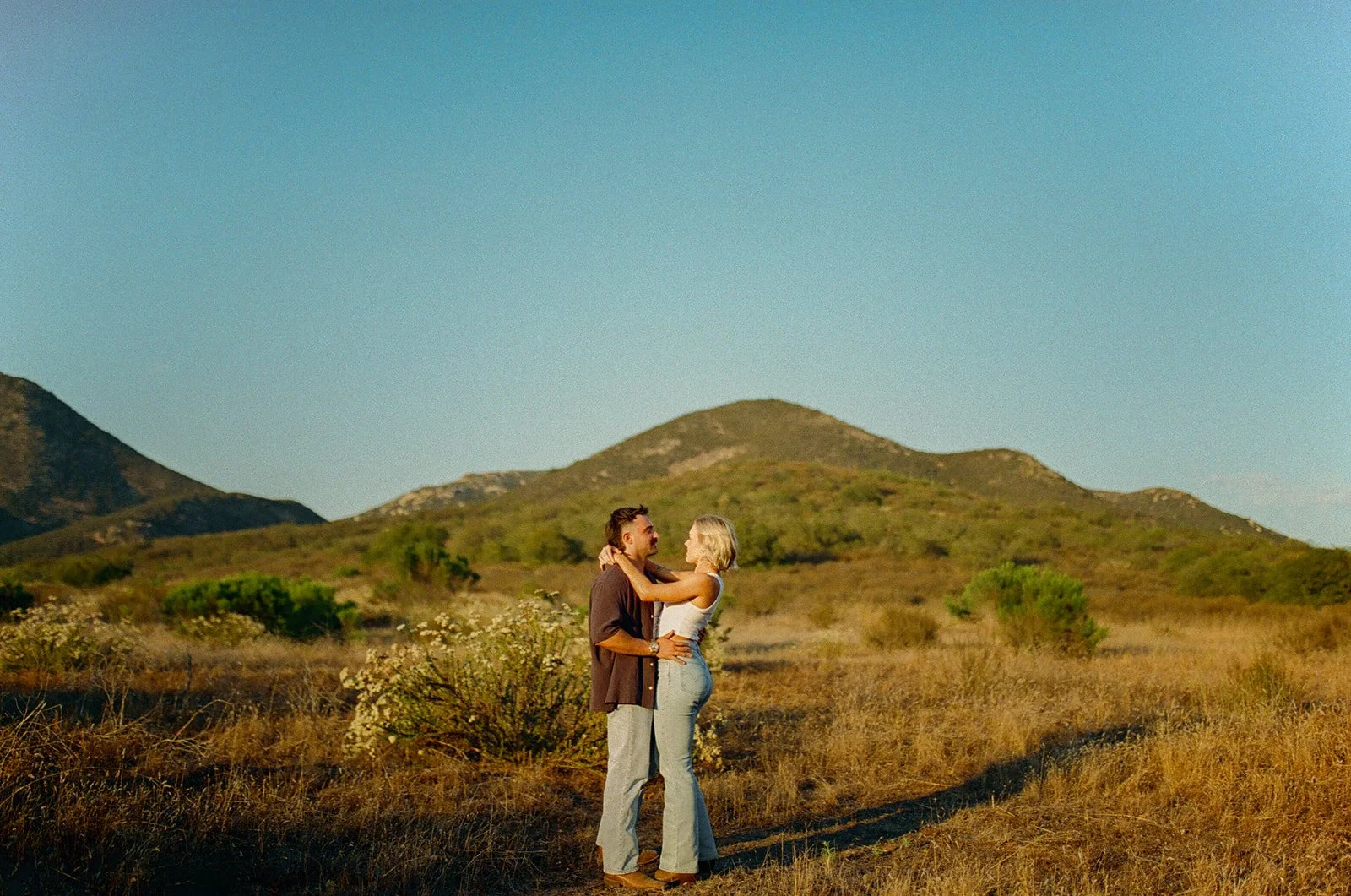 Wide landscape shot of the couple embracing in the middle of a grassy mountain field during golden hour.