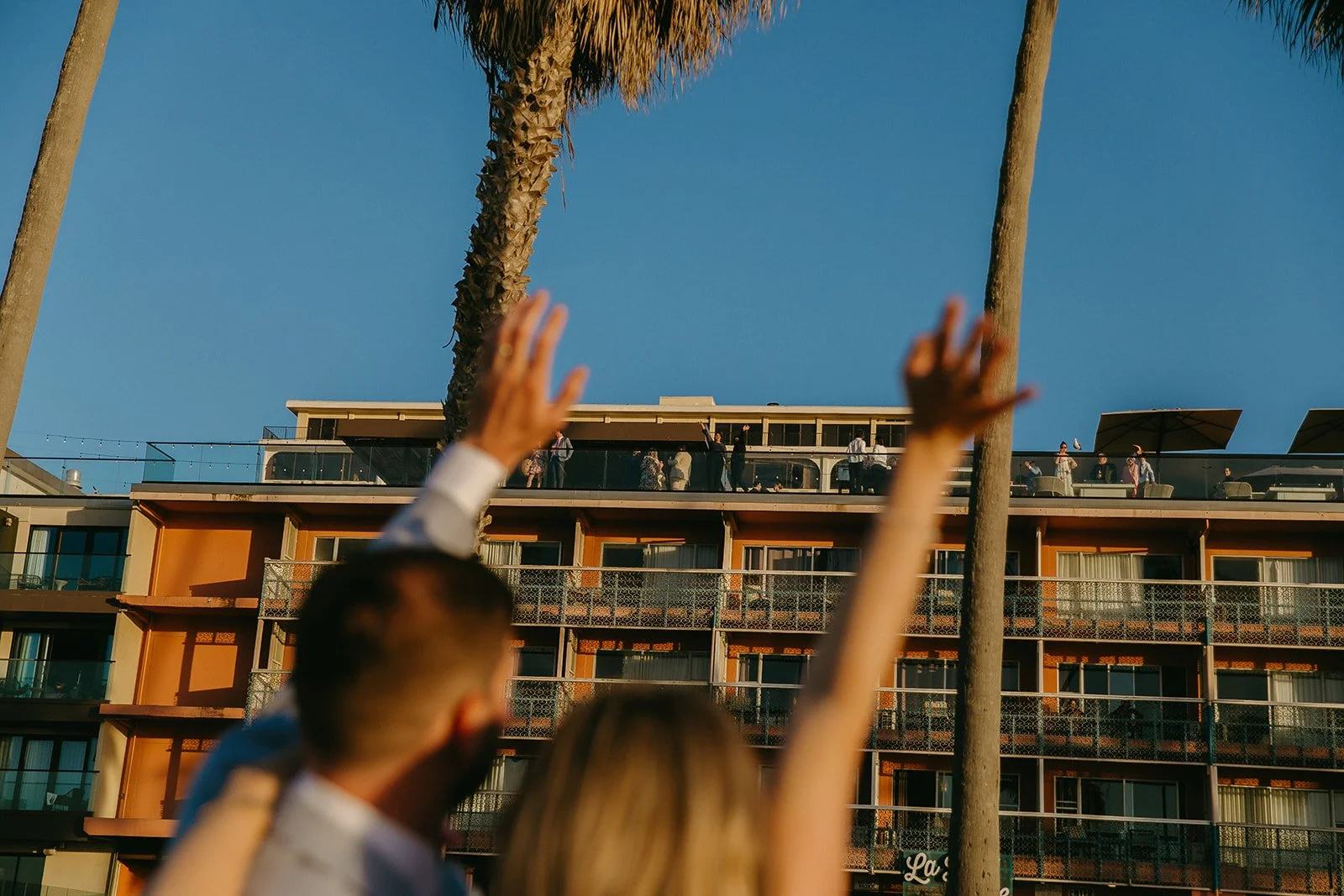 A couple waving at wedding guests on a rooftop from below