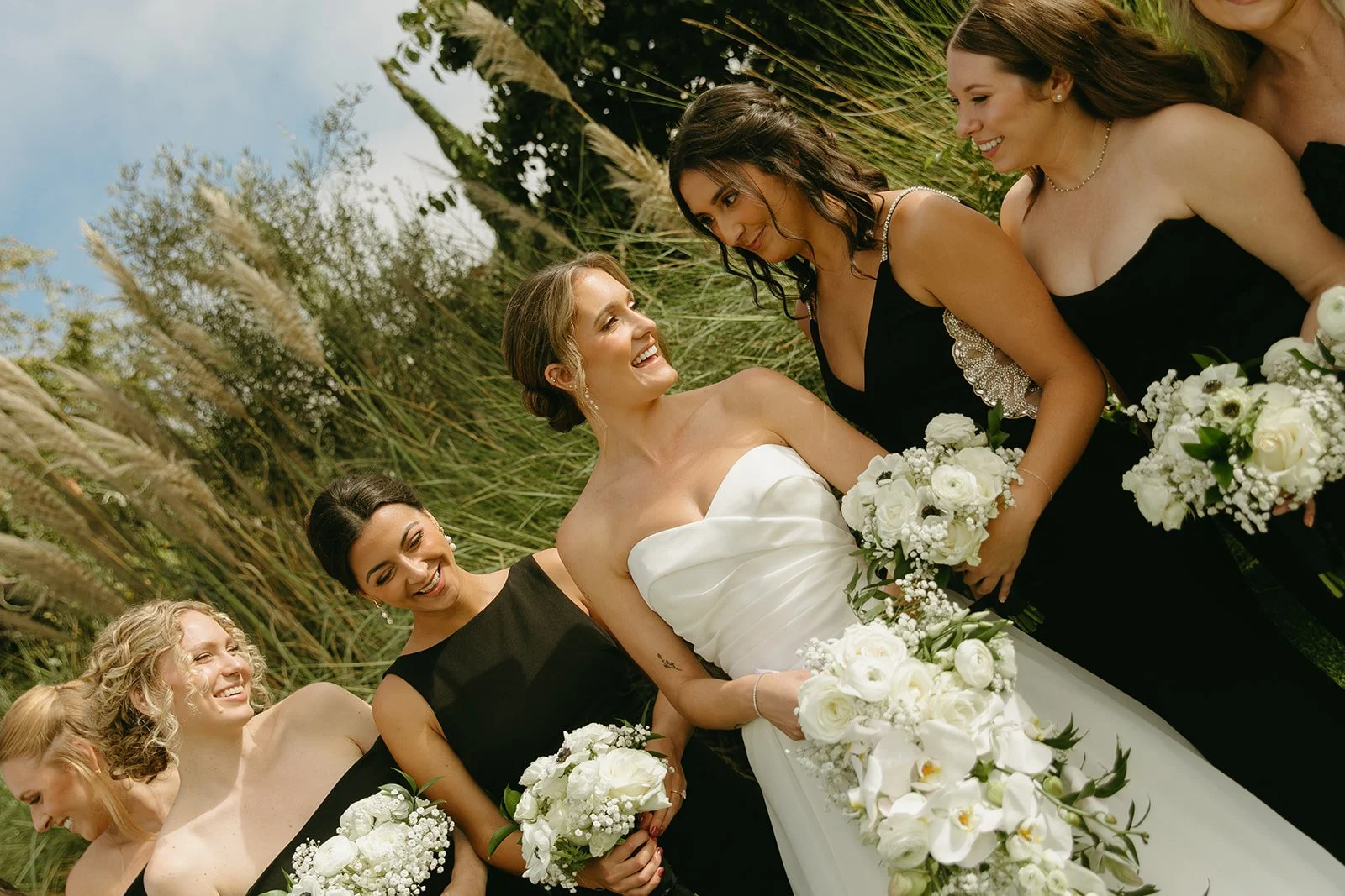 Bride standing with her bridesmaids in black dresses, laughing together while holding white floral bouquets during portraits.