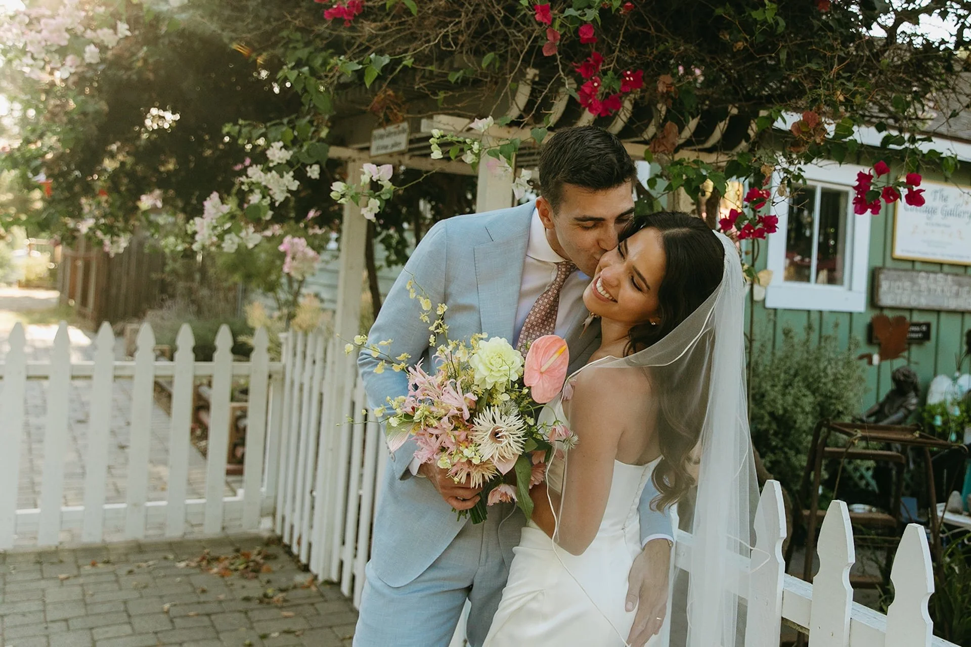 The groom kissing the brides cheek as they stand in front of some blooming flowers during their bride and groom portraits