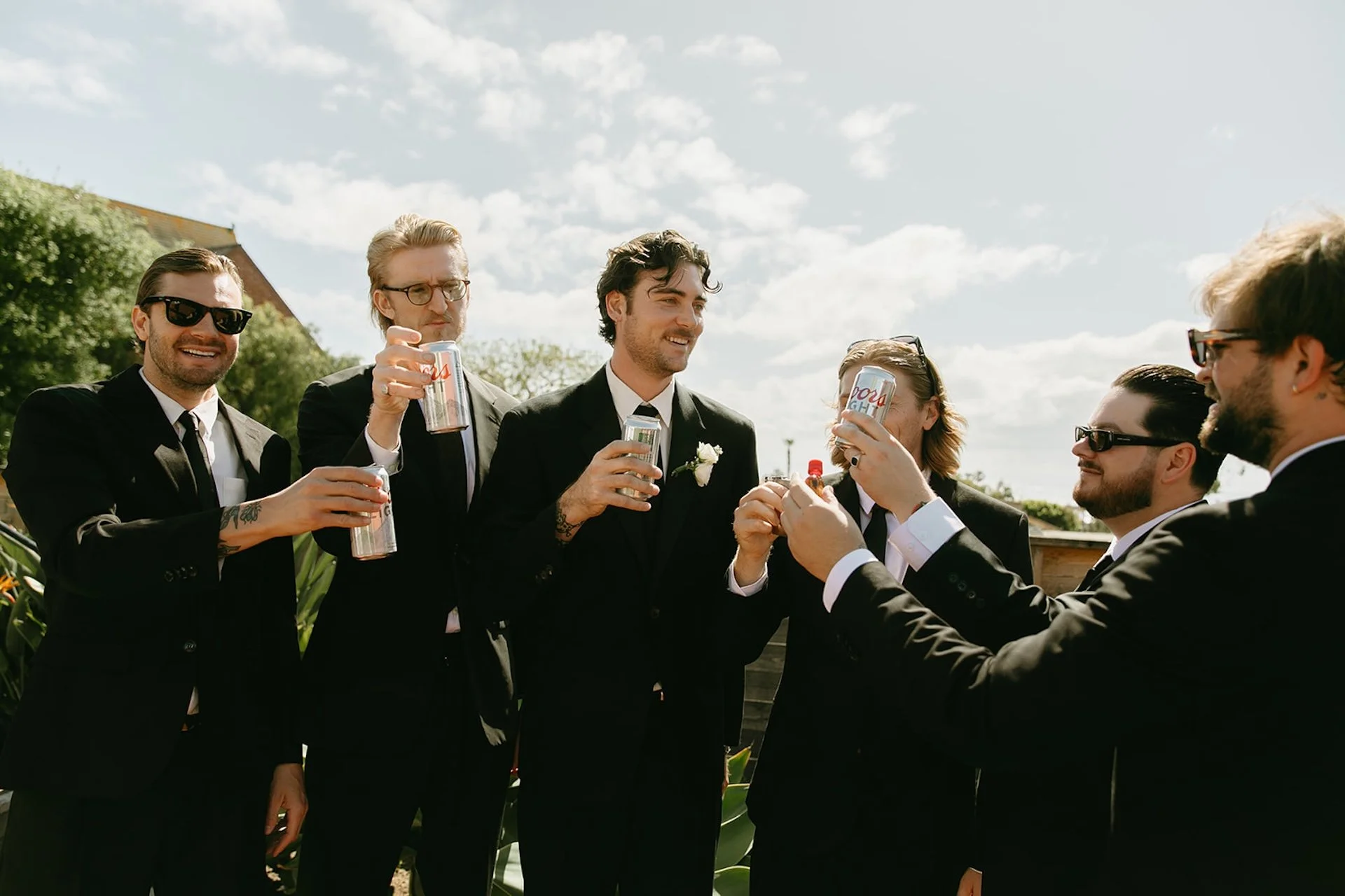 Groom celebrating with groomsmen while they toast drinks together outdoors in black suits.