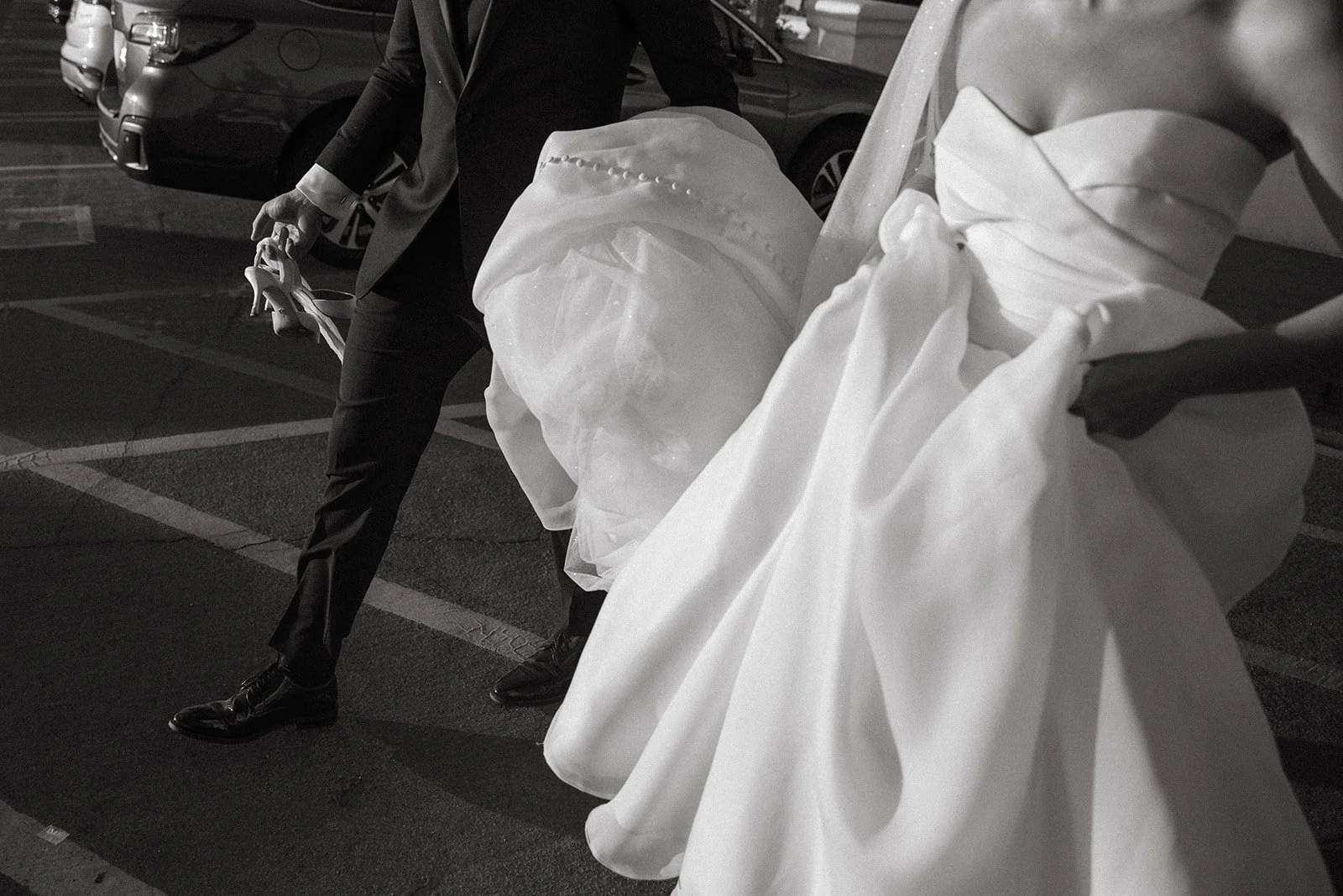 Black and white candid moment of the bride and groom walking together, holding their wedding attire as they move through a parking area.