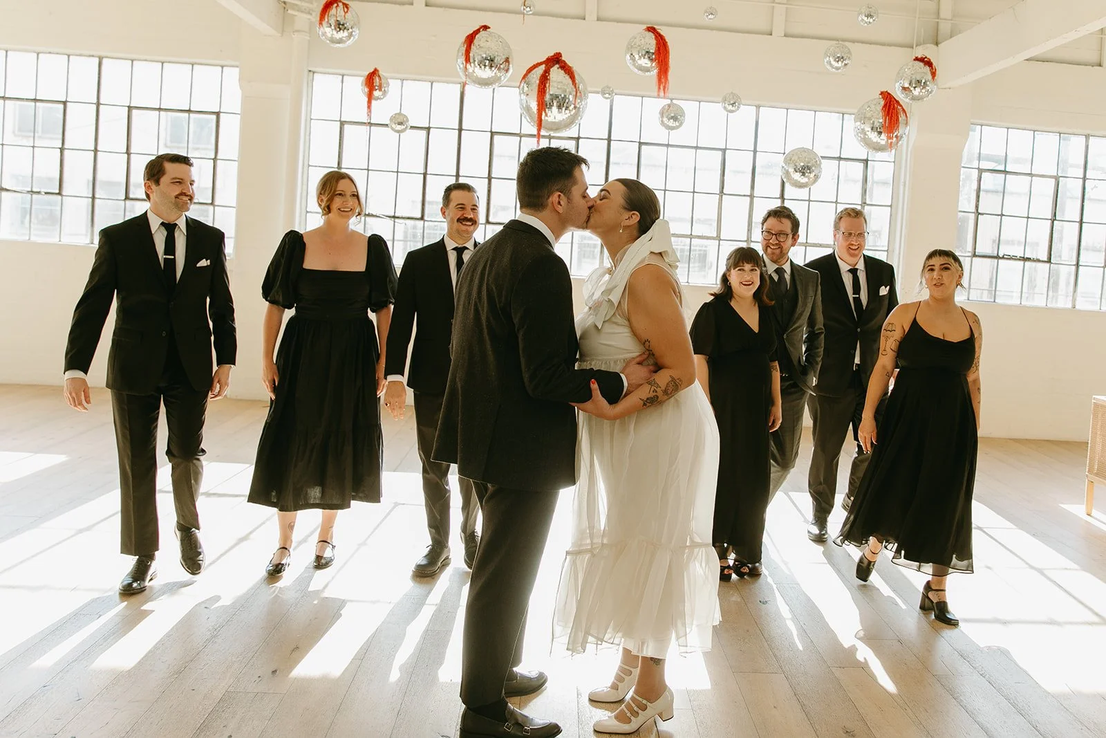 The couple sharing a kiss surrounded by their wedding party inside The Revery LA, with disco balls and sunlight filling the space.