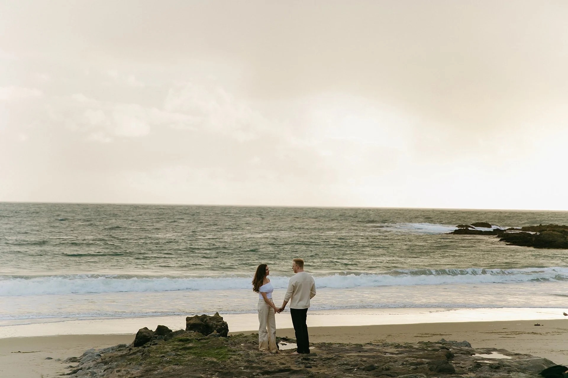 A couple holding hands while standing on a rocky outcrop above the beach, facing the ocean as waves crash along the shore.