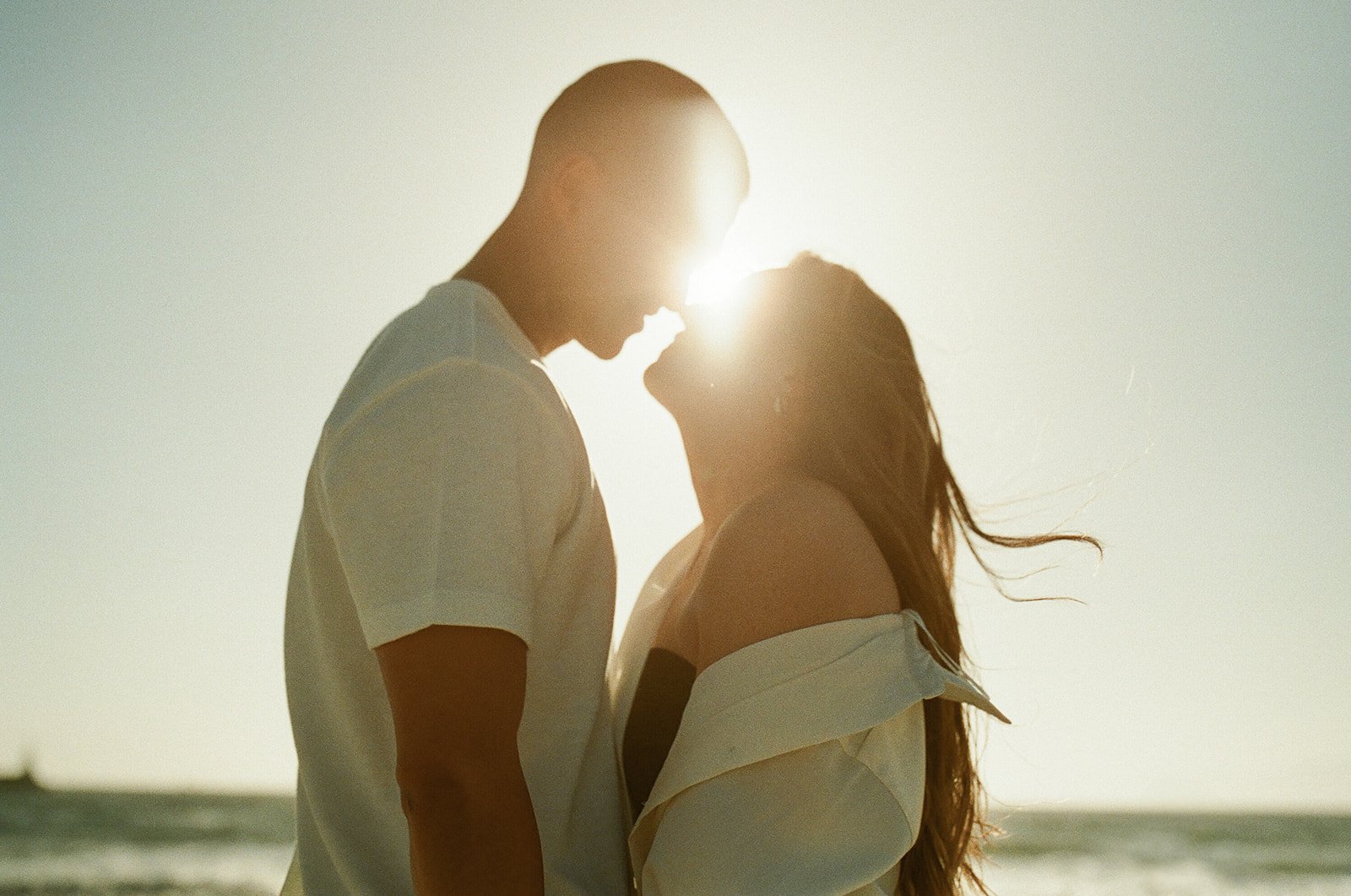 A romantic silhouette of a couple standing forehead to forehead at the beach with the sun setting behind them.