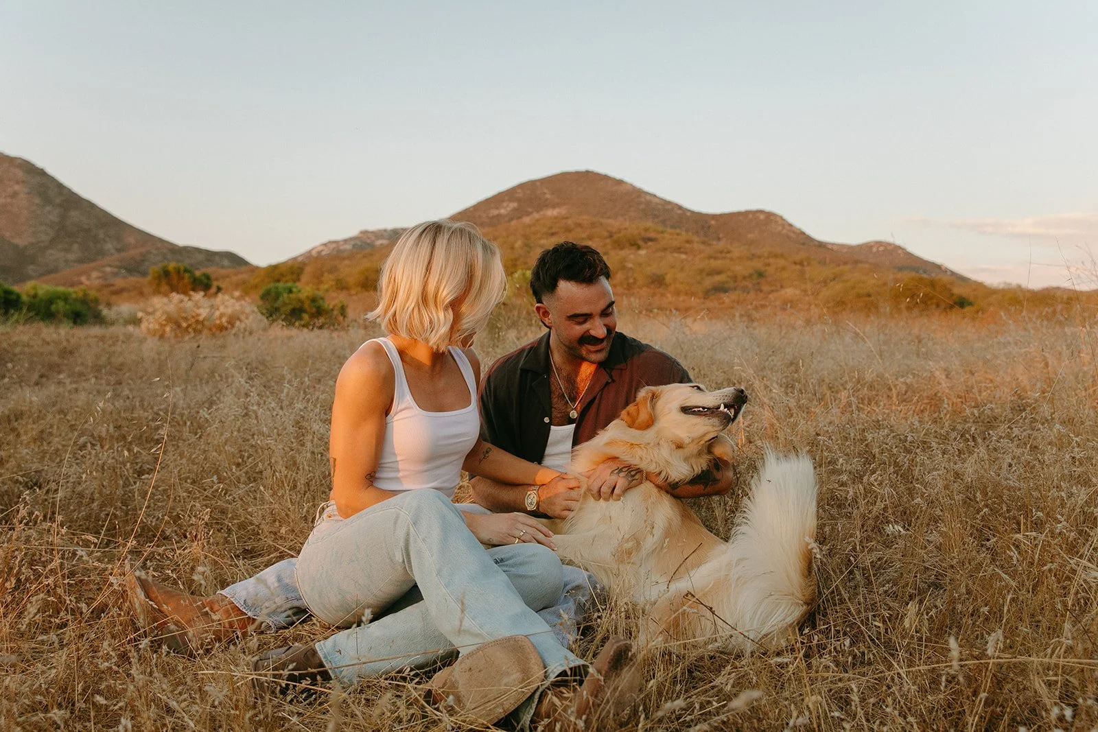 Black and white portrait of the couple smiling while holding their golden retriever together in an open meadow.