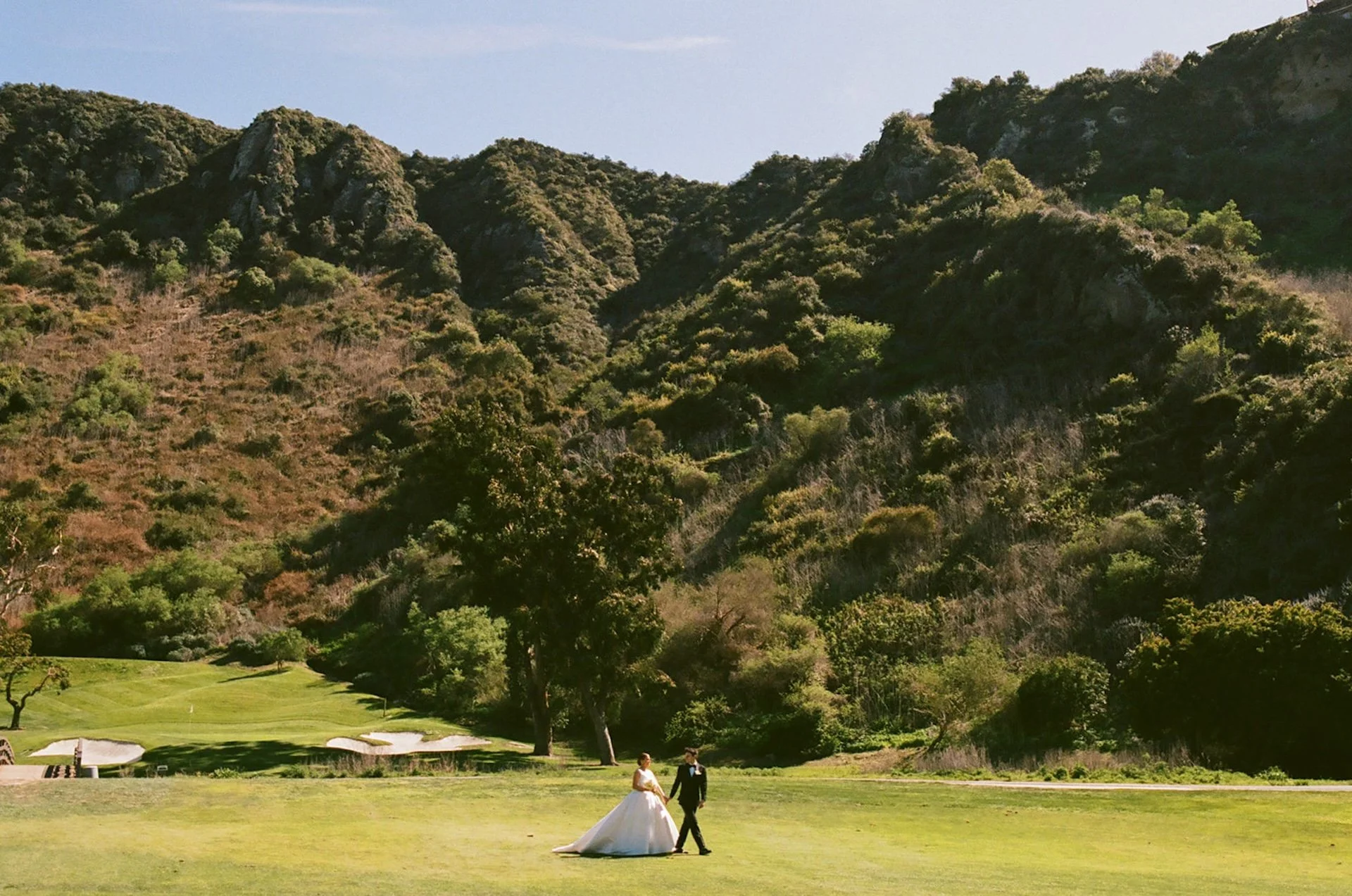 Bride and groom walking across the golf course with canyon hills behind them during a romantic outdoor wedding portrait at The Ranch at Laguna Beach wedding venue.