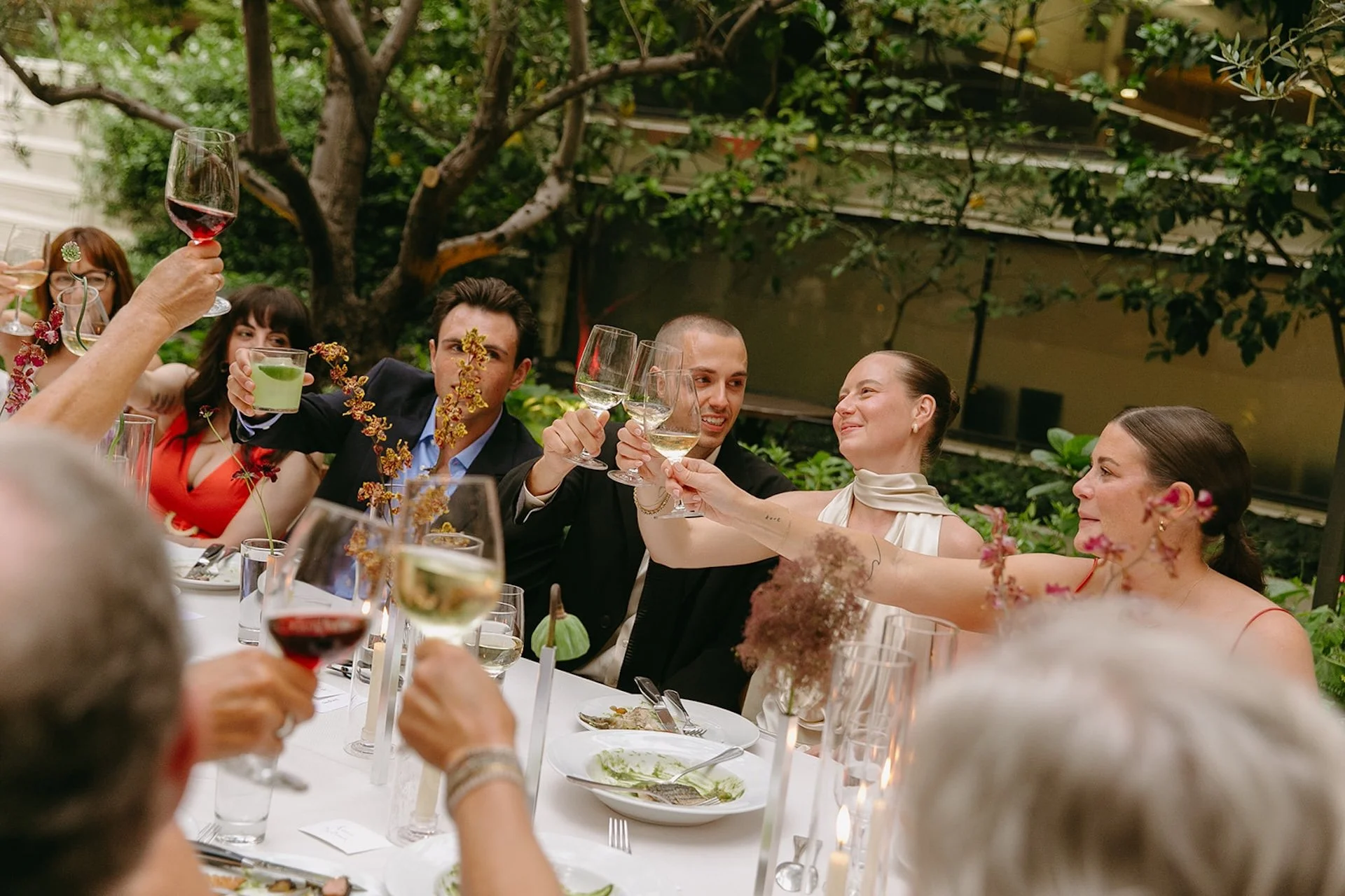 The couple clinking wine glasses with guests during dinner, smiling and seated at a long banquet table surrounded by greenery.