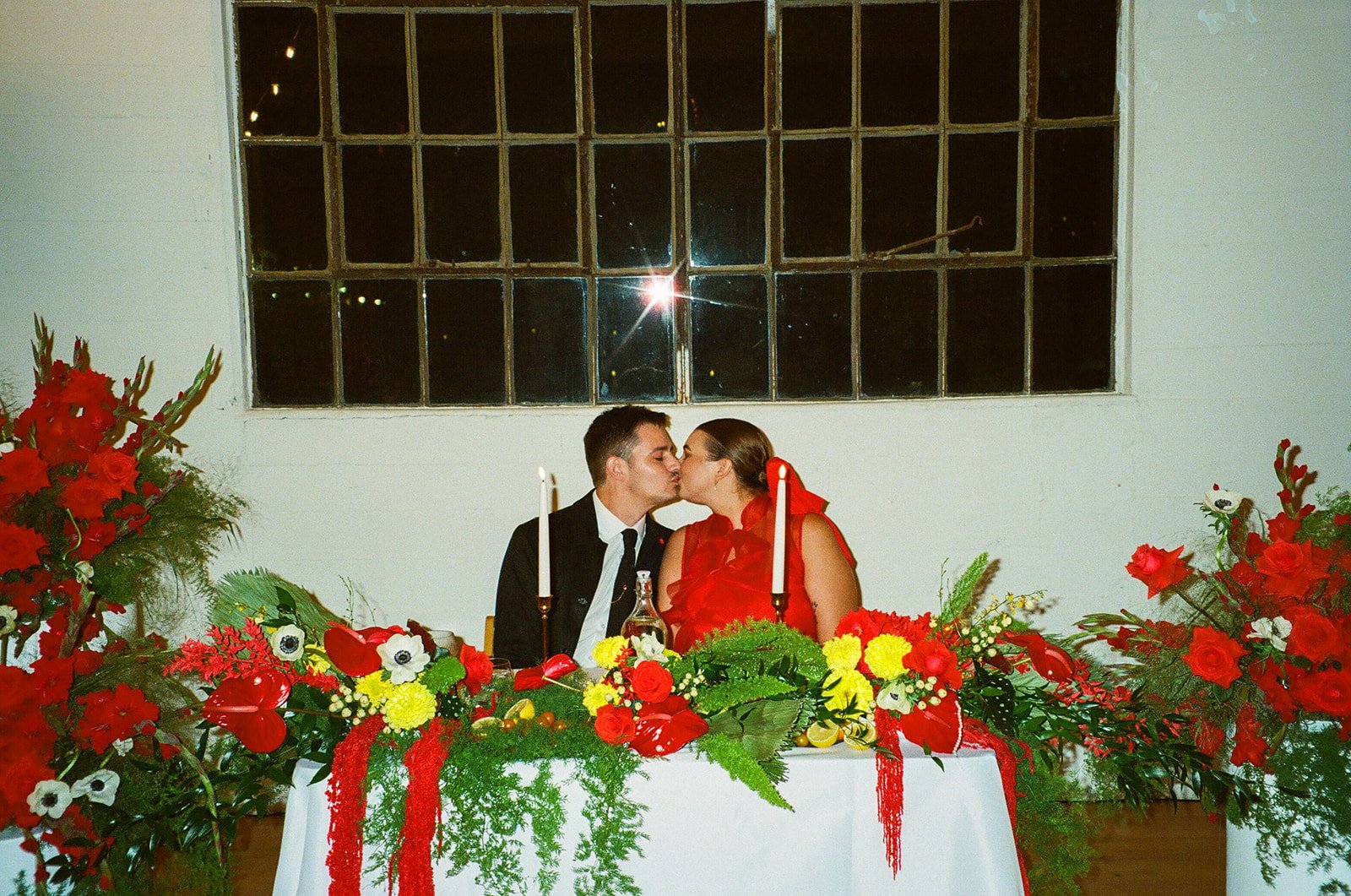 The couple seated at their sweetheart table at The Revery LA, surrounded by bold red florals, candles, and colorful tablescape details.