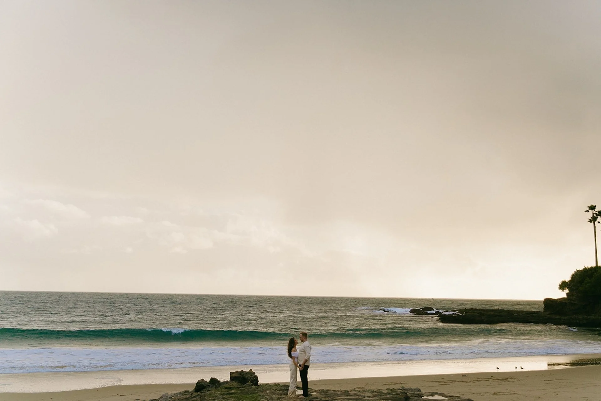 Distant view of the couple standing alone on the beach with waves rolling in, photographed as laguna beach engagement photos at shaws cove laguna beach under a moody sky.