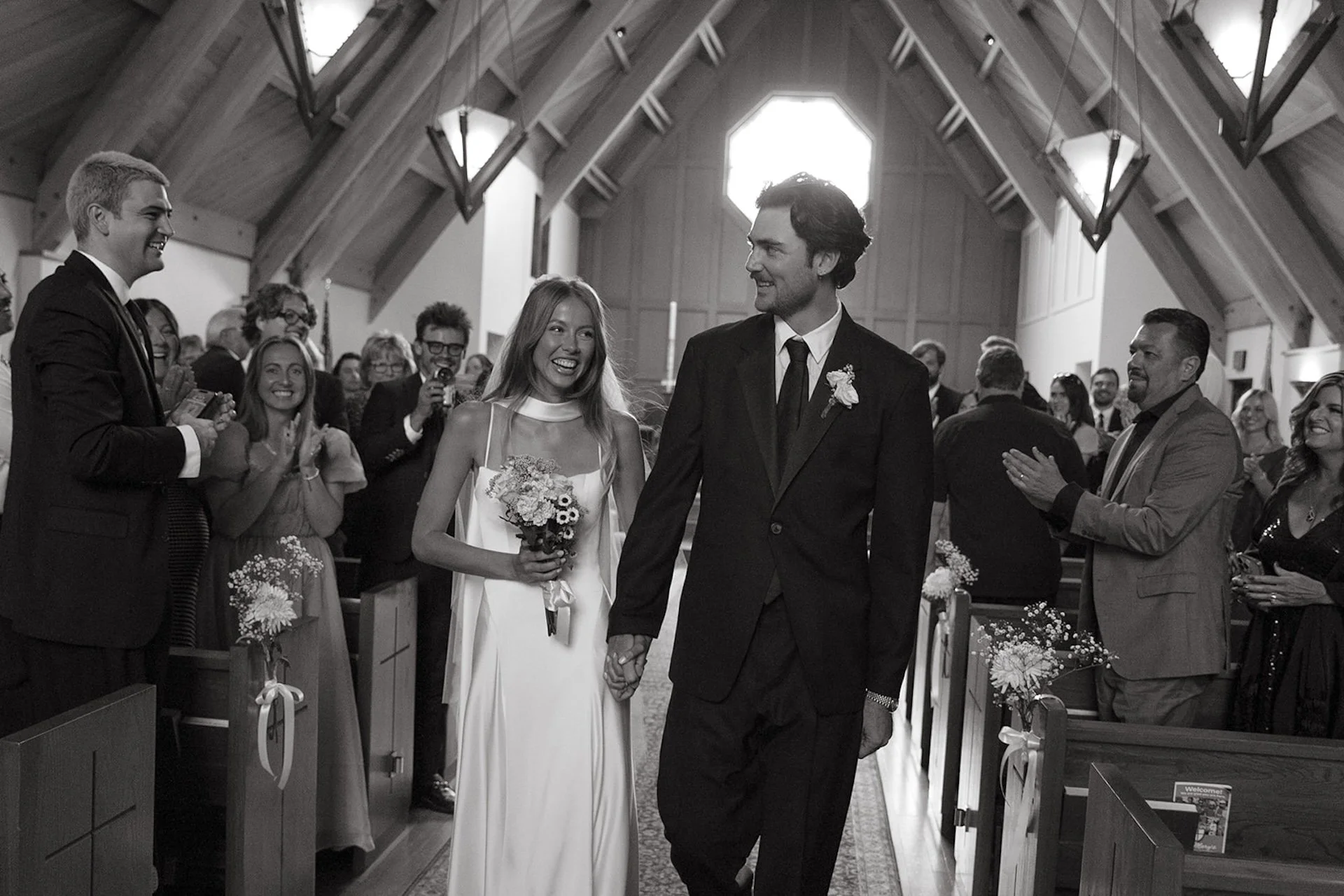 Newly married couple walking hand in hand down the aisle while guests smile and applaud inside the chapel.