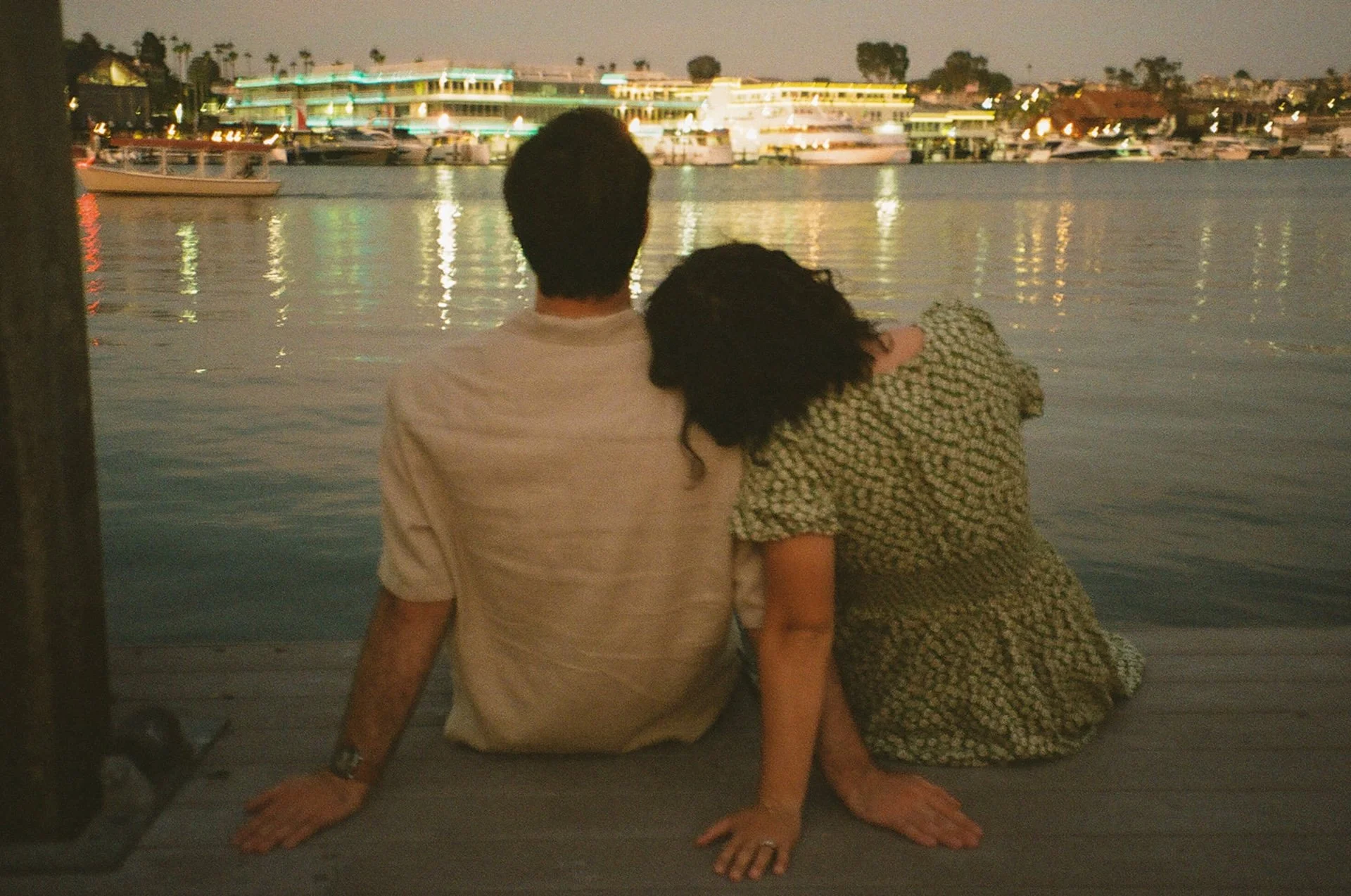 Nighttime candid of a couple sitting on a dock by the water, laughing and cuddling under city lights reflecting across the harbor.