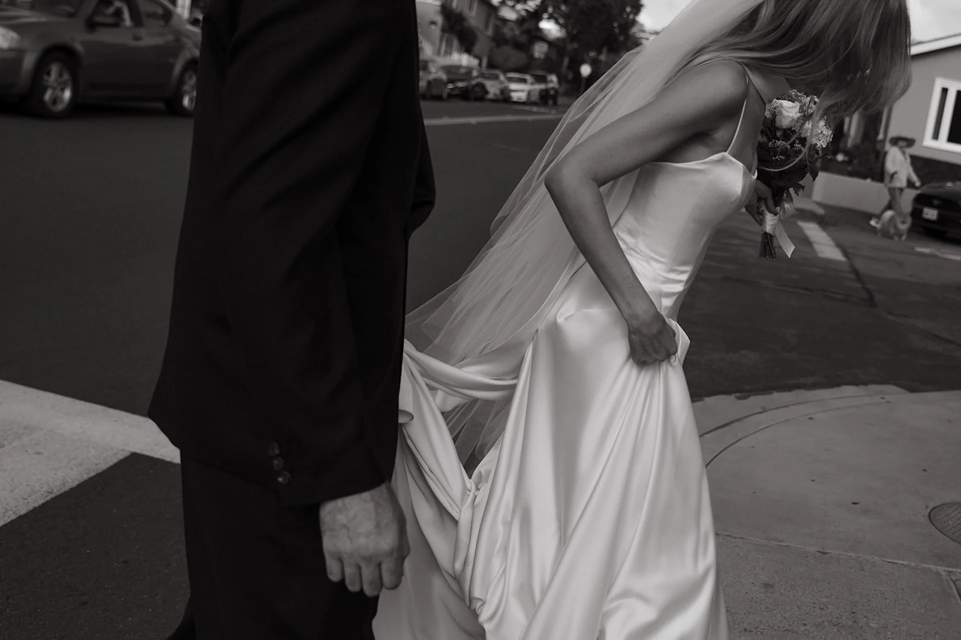 Bride lifting her satin wedding dress while walking across a quiet Laguna Beach street with her bouquet and veil flowing behind her.