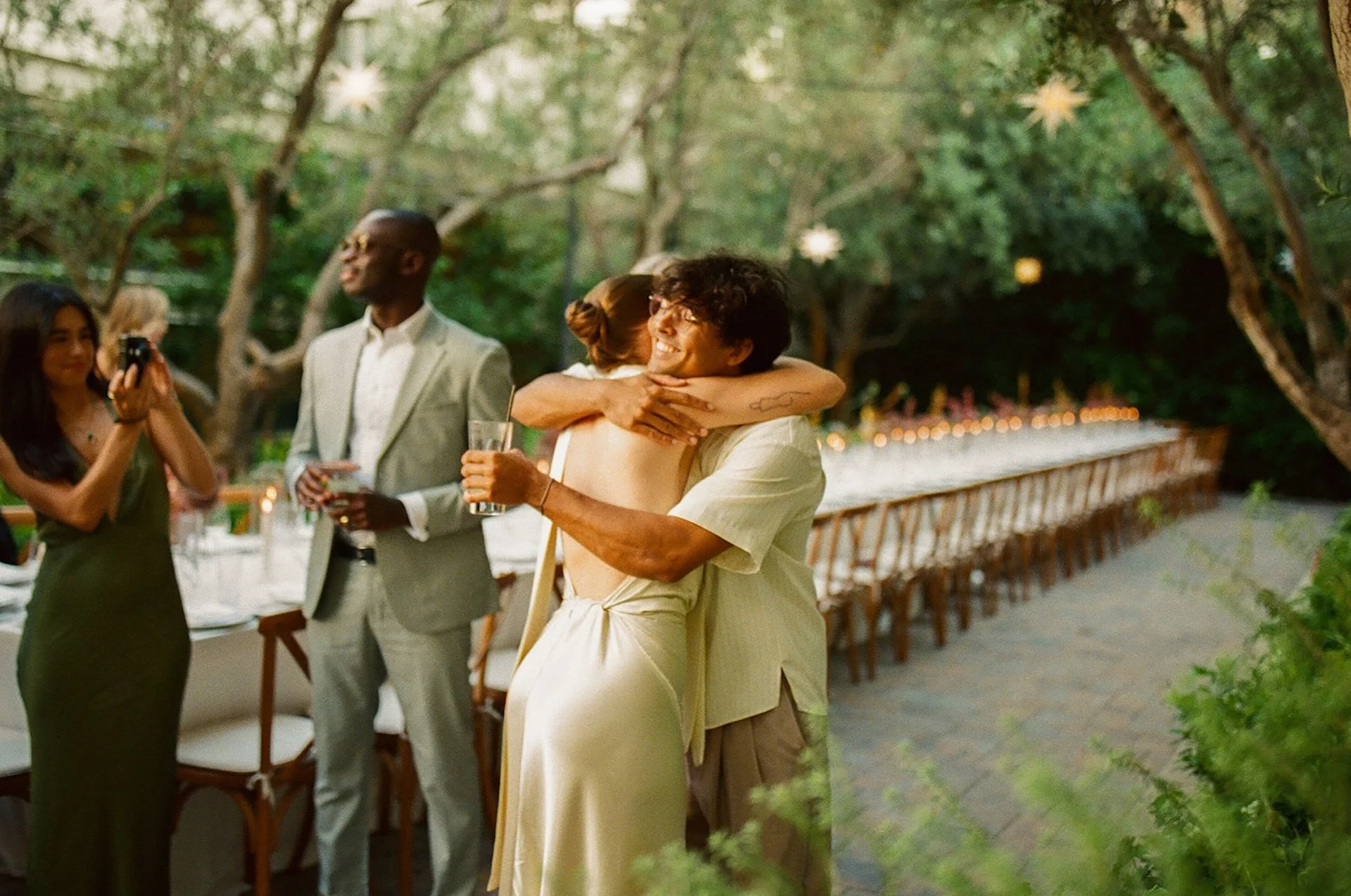 The couple hugging friends and guests during the reception, with blurred motion and greenery creating a lively, documentary-style wedding scene.