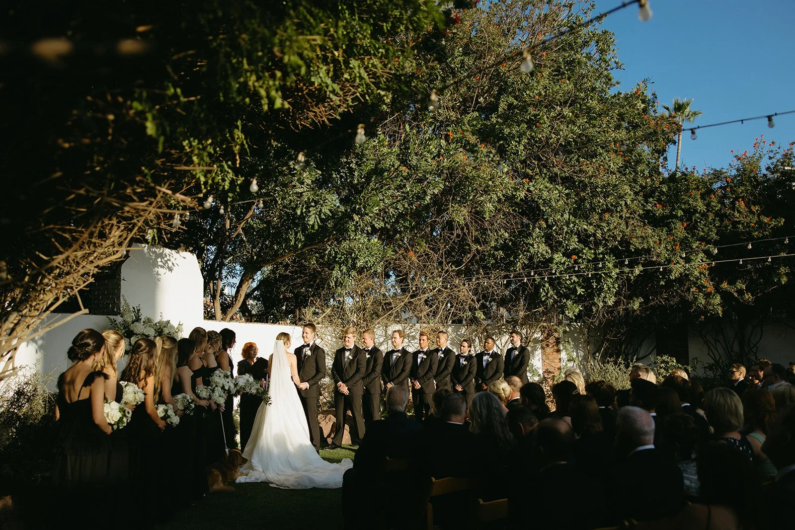 Wide ceremony view with the bride and groom standing at the altar beneath string lights at one of the most romantic San Clemente Wedding Venues, surrounded by lush greenery and guests.