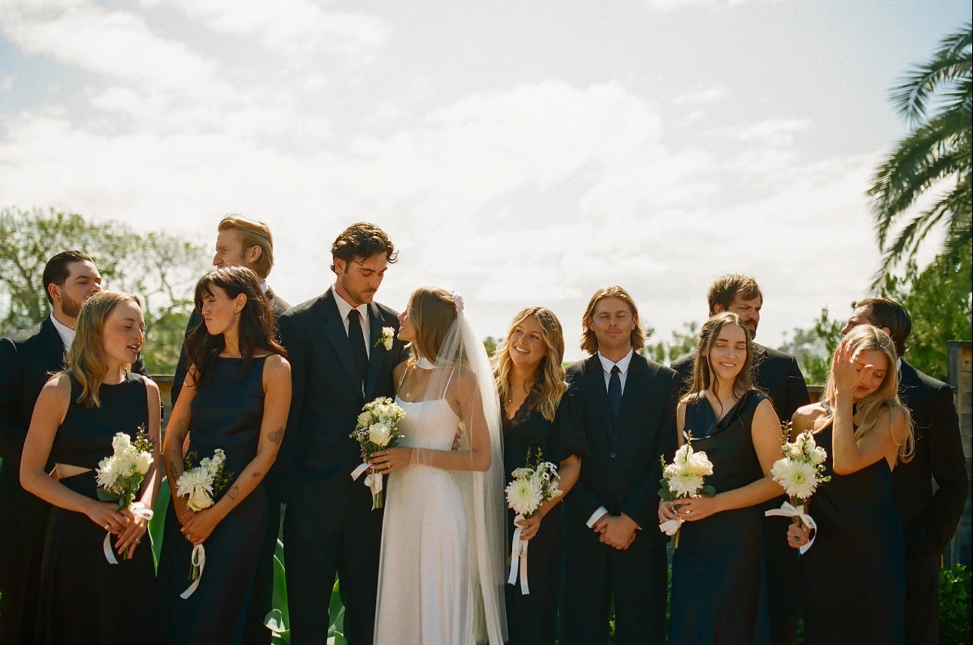 Wedding party standing together outdoors in black attire with white florals during a sunny coastal ceremony portrait.
