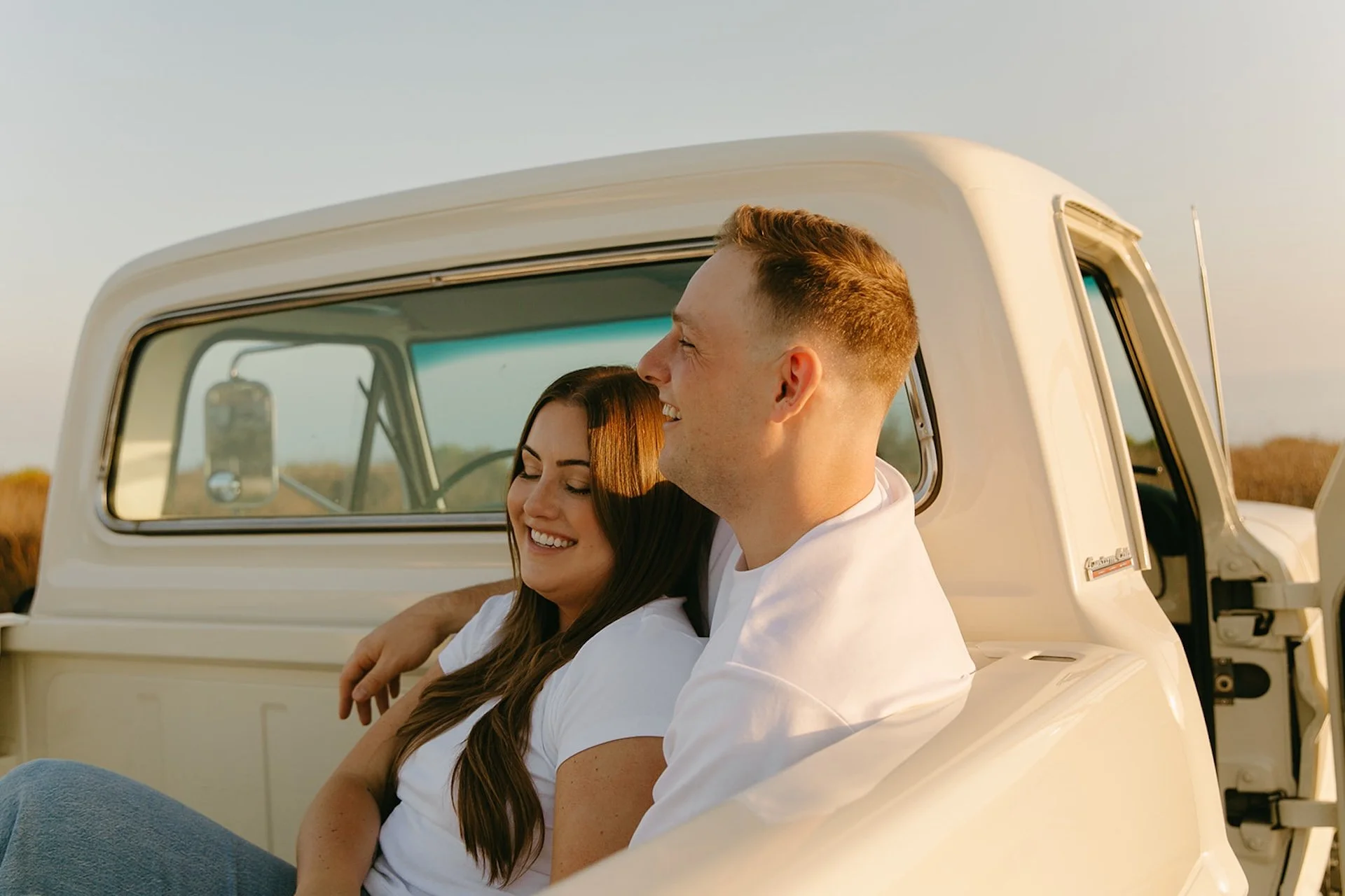 Beach Engagement Photo Ideas of a couple sitting in the bed of a vintage truck at sunset, holding each other and laughing with tall grass and golden light behind them.