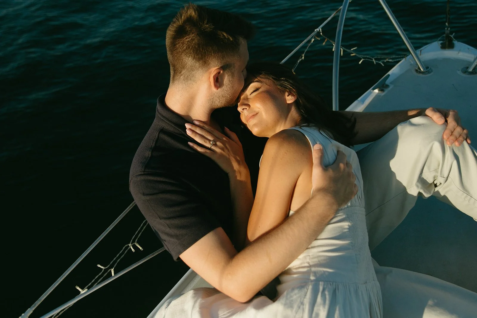 Couple snuggling close while seated on a sailboat, sunset sailboat engagement photos with warm evening light.