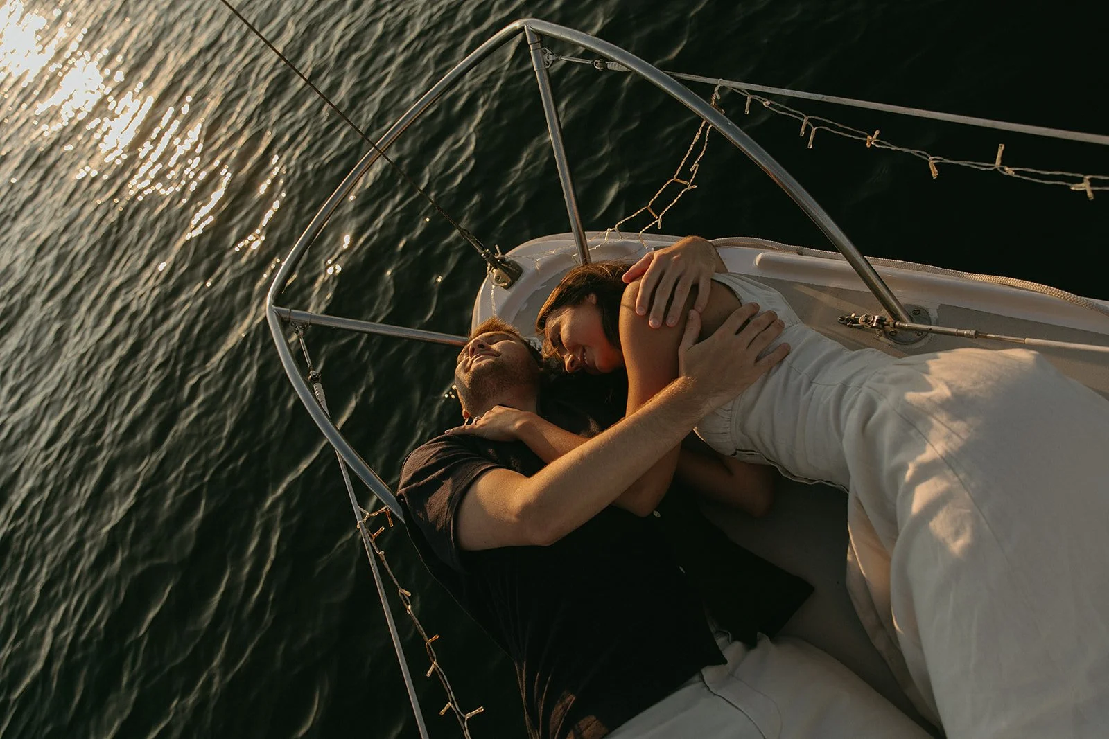 Couple laying down on the front of the boat during their sailboat engagement photos with the water in the background as the sun sets.
