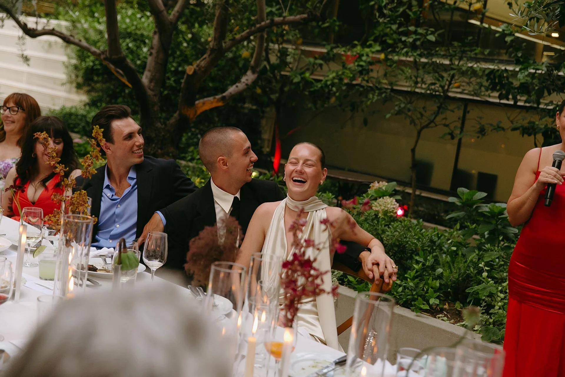 A candid color photo of the couple laughing during speeches at the dinner table, with floral centerpieces and candles creating a warm, intimate atmosphere.