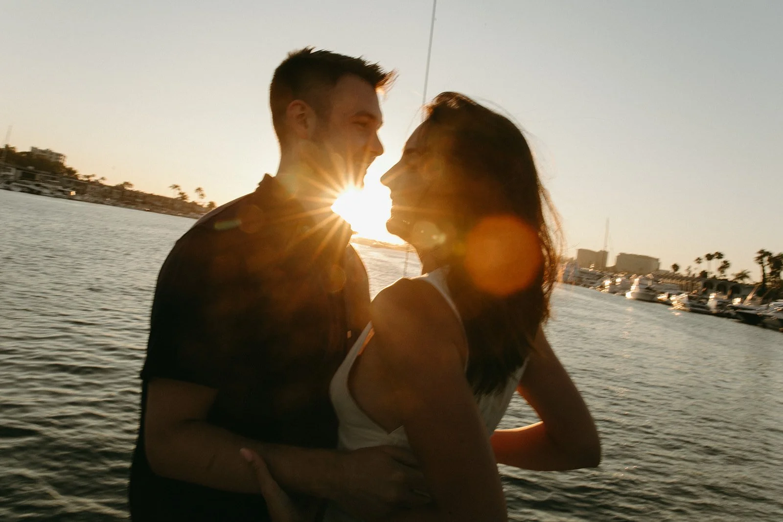 Playful couple laughing as the sun sets over the water, candid summer sailboat engagement photos.