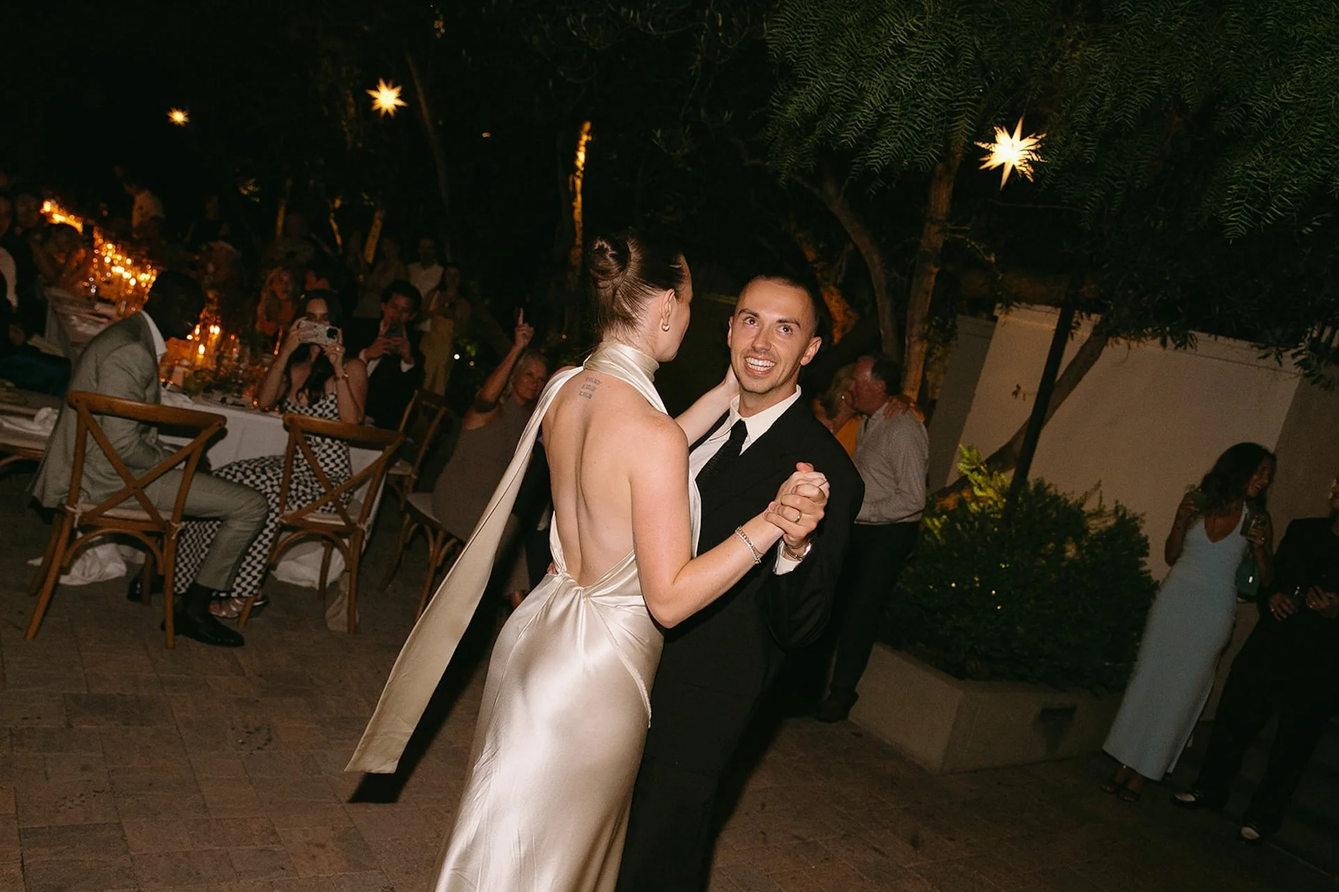 The newlyweds slow dancing during their first dance, with the groom in a black suit and the bride in a satin halter gown, holding each other closely in a dimly lit outdoor setting.