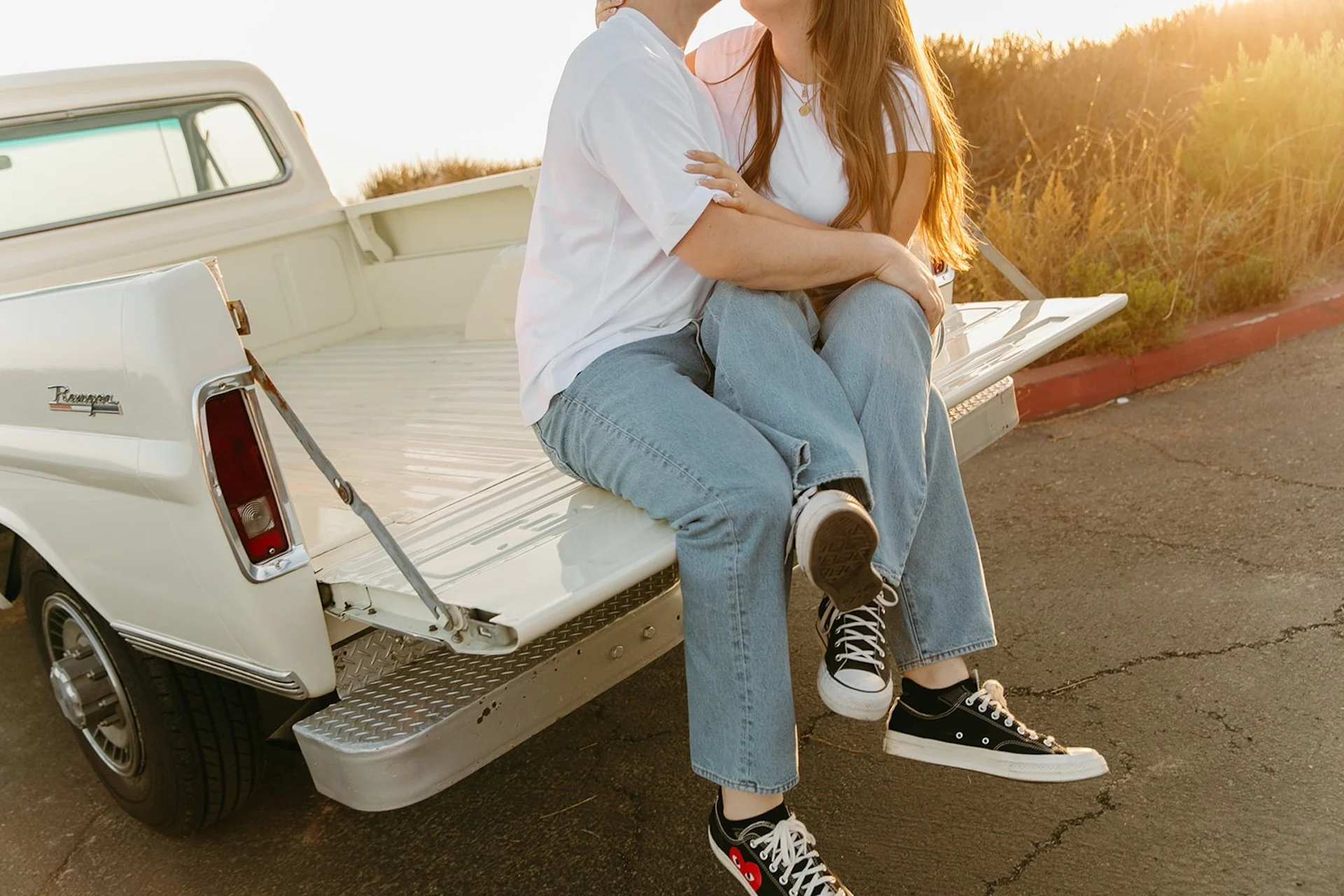 Beach Engagement Photo Ideas of a close-up of a couple sitting on the tailgate of a vintage truck, focusing on their intertwined legs in denim and black Converse sneakers with warm sunset light.