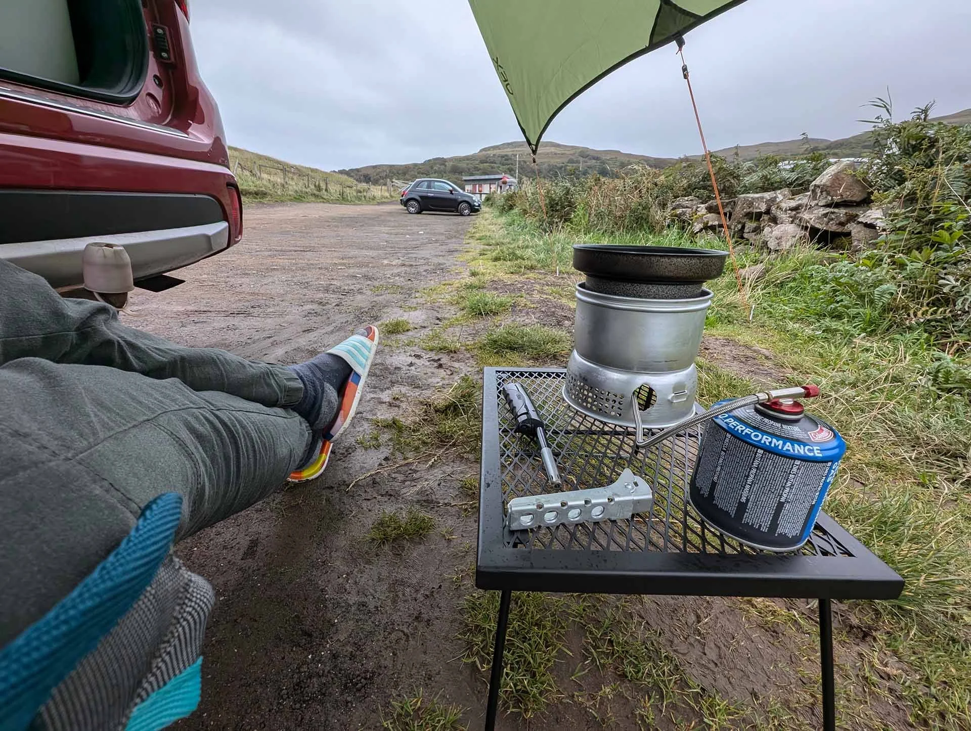 A person (whose legs you can see on the left-hand side of the image) sits next to a Trangia with a gas canister attached, waiting for their dinner to cook. They sit under a tarpaulin that is stretched over the back of their car.