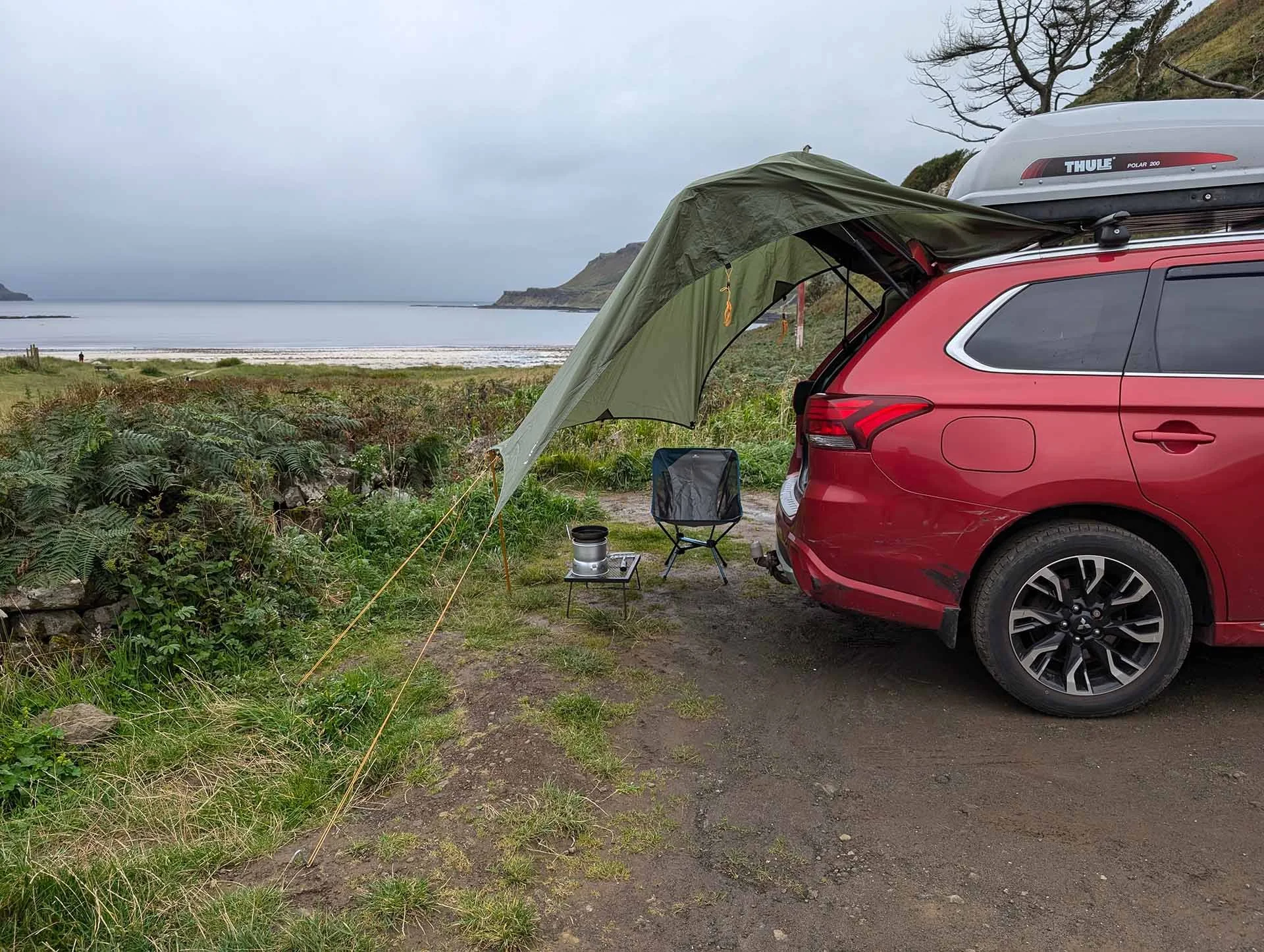 The back end of a red Mitsubishi Outlander with the boot open. Stretched across the boot is a tarp which creates a living space that houses a camping stove and a chair.