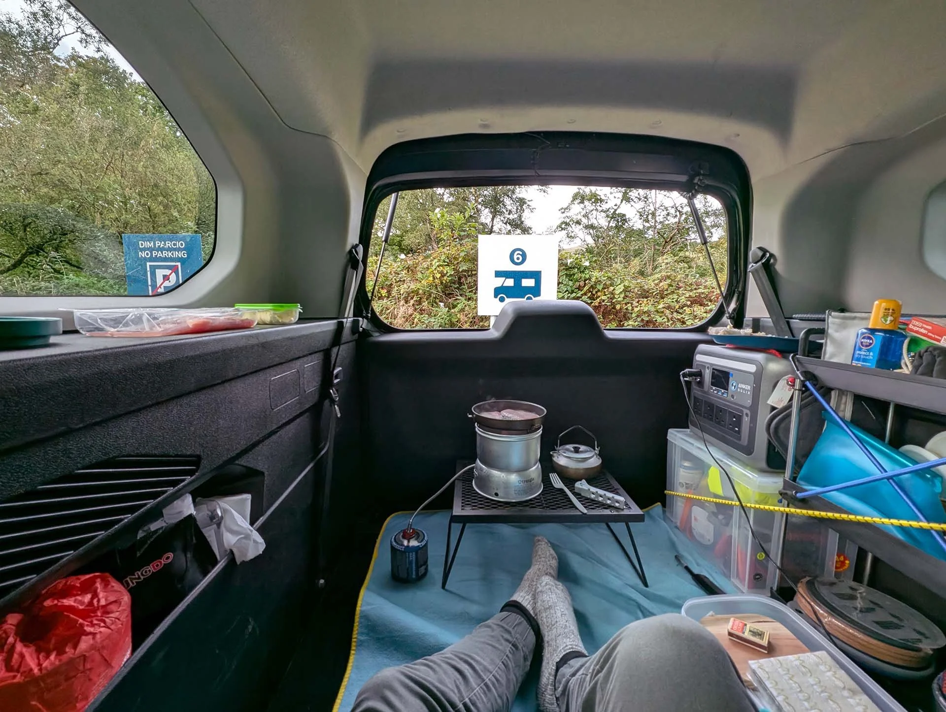 A Trangia stove stands on a small BBQ stand with a pan of frying bacon on top. In the foreground are a pair of legs as the person sits, waiting for the bacon to cook.