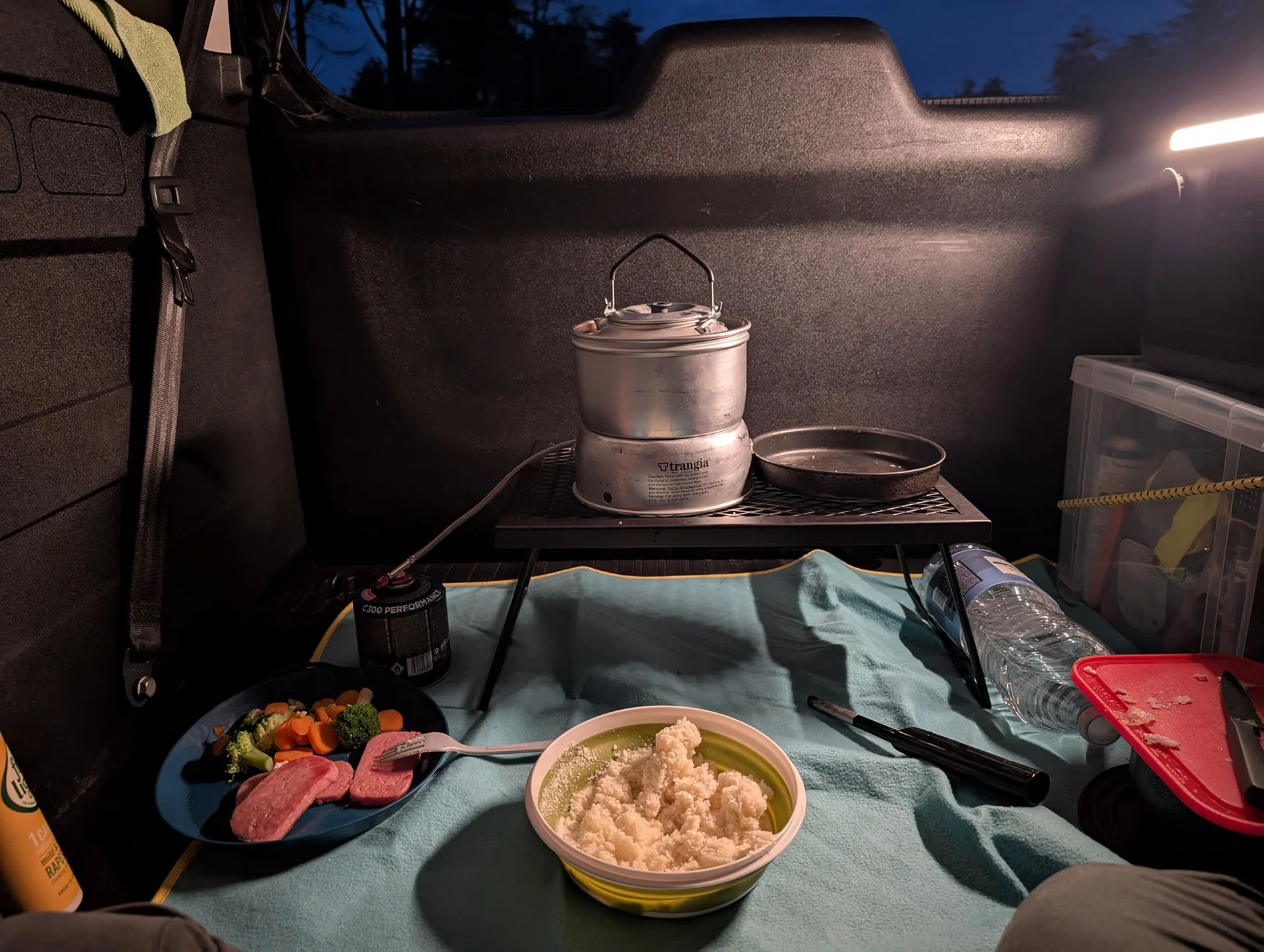 A Trangia stands on a small BBQ stand inside a Citroen Berlingo. In front of the stove is a bowl containing instant mashed potato and a plate with cooked Spam and vegetables.
