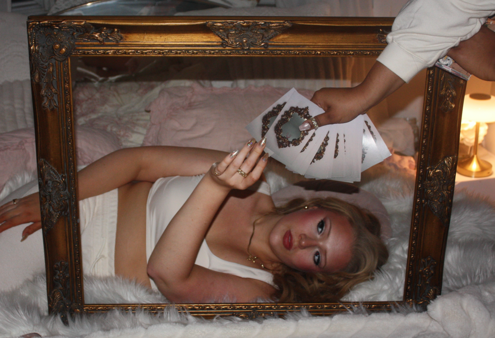 Woman lying on bed with head on pillow, holding a set of nail art stickers. Reflected in a decorative mirror in a cozy, well-lit bedroom with pink bedding and a bedside lamp.