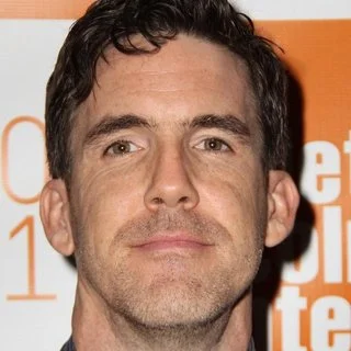 Close-up of a man's face with short dark hair and light skin, standing against an orange and white background.