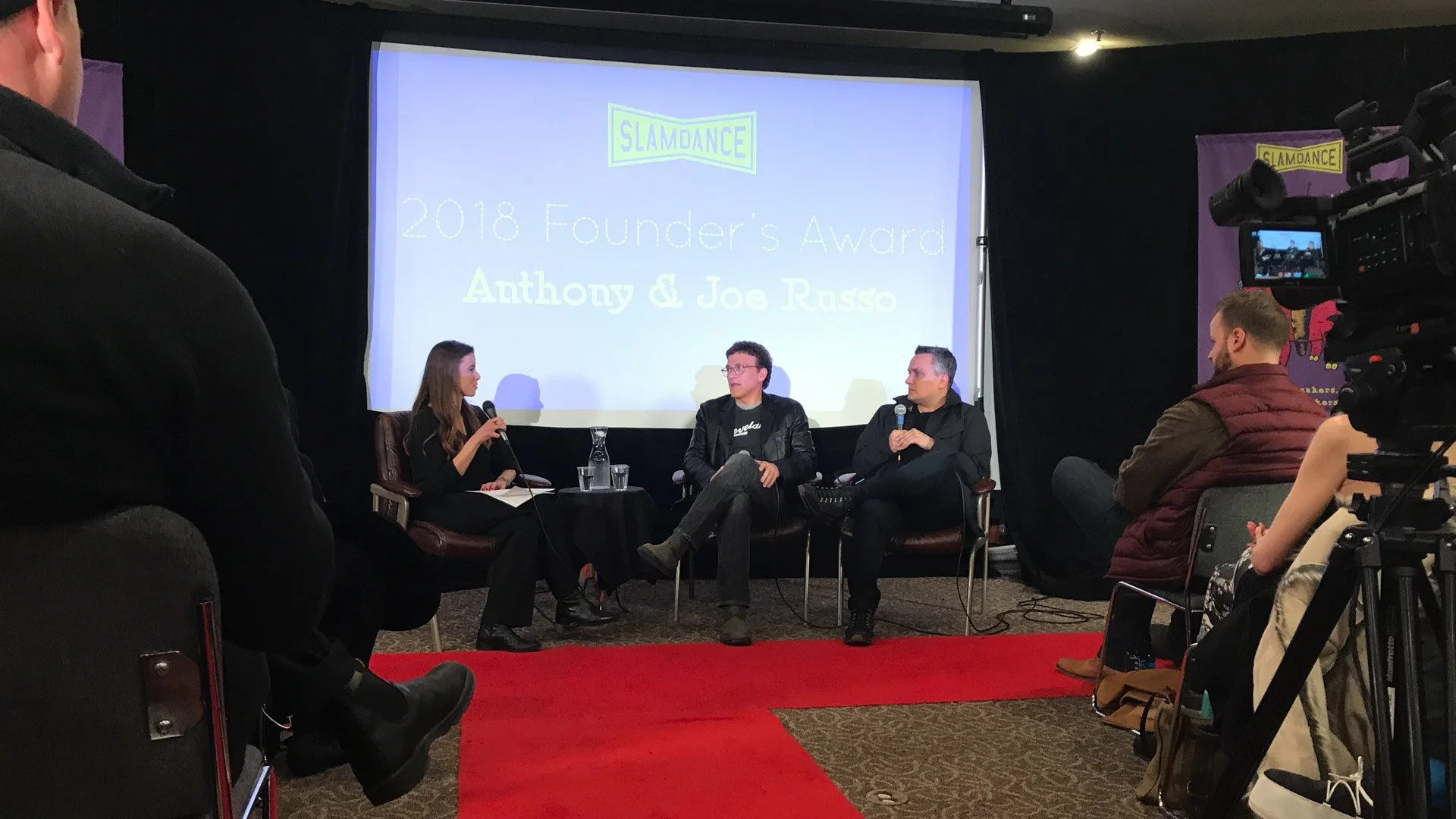 Panel discussion moderated by Sandra Bertalanffy at the Slamdance 2018 Founder's Award with Anthony & Joe Russo seated in front of a large screen displaying the event's name and guests, with an audience and camera capturing the scene.