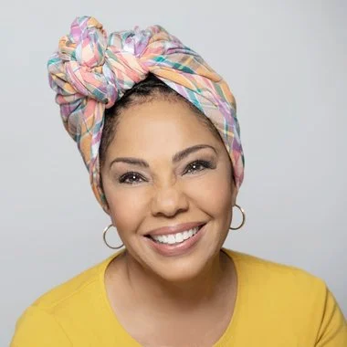 Smiling woman with a colorful headwrap and gold hoop earrings, wearing a yellow top.