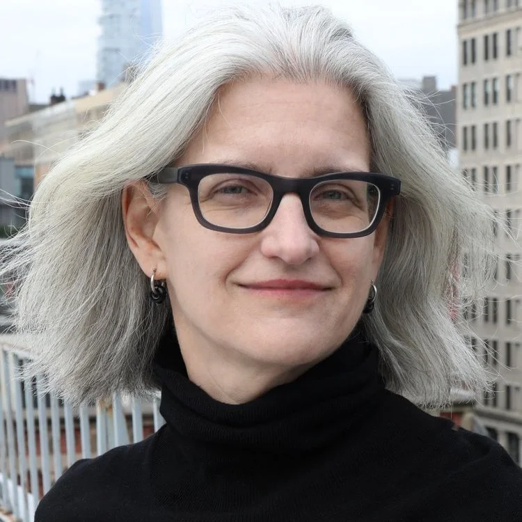 A woman with gray hair, black glasses, and earrings, wearing a black turtleneck, standing outdoors in an urban setting with buildings in the background.